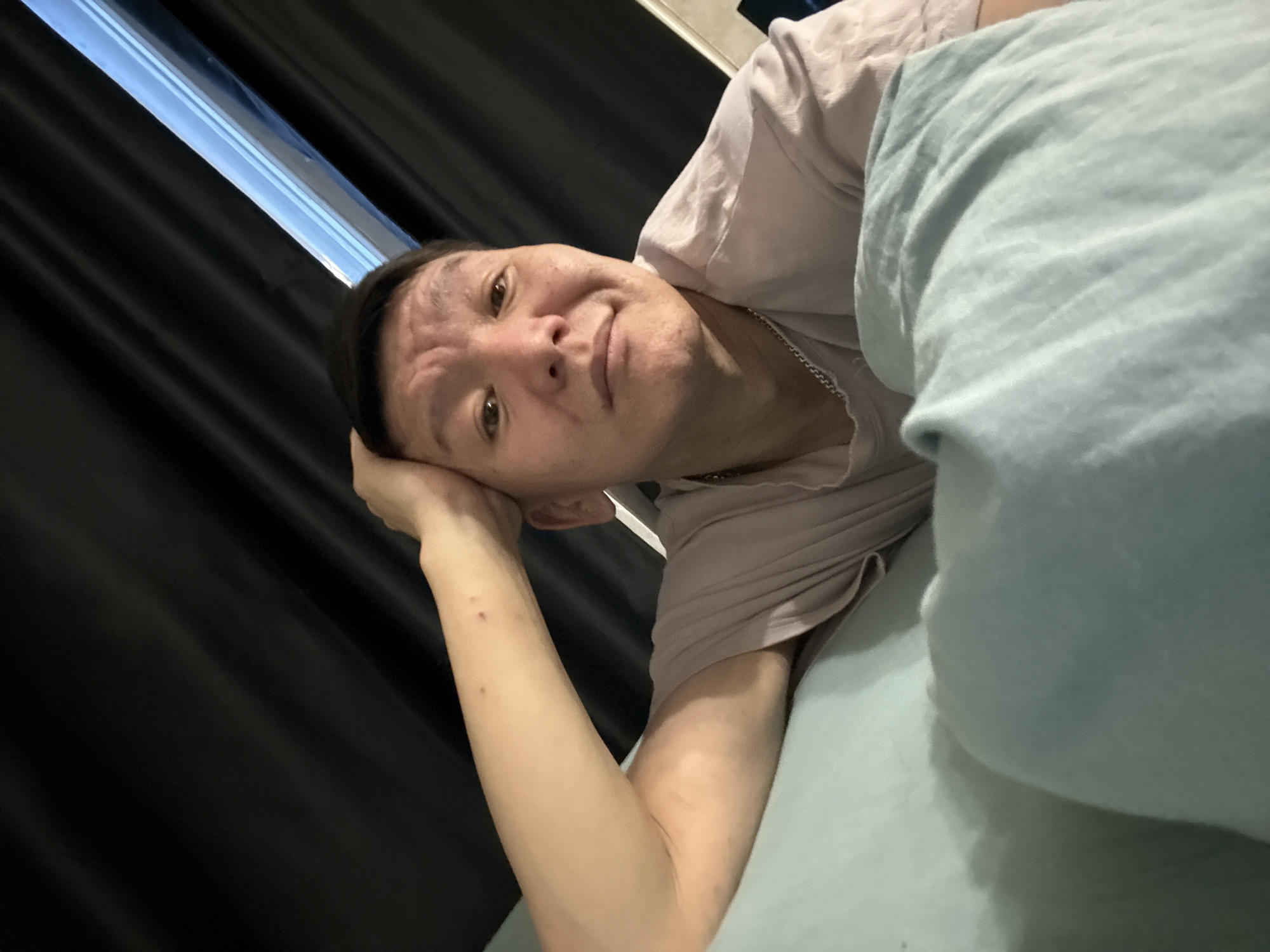 The image is a selfie taken indoors. A person is lying down, with their head resting on their hand. They are wearing a light purple t-shirt and a silver chain necklace. The background consists of dark curtains and a bright light source coming from a window or skylight. The person's face is partially obscured by their hand and they appear to be looking at the camera with a neutral expression. The overall impression is of a casual, relaxed setting, possibly a bedroom. The location is not specified in the image, and therefore the city of Khimki, Russia, cannot be confirmed.