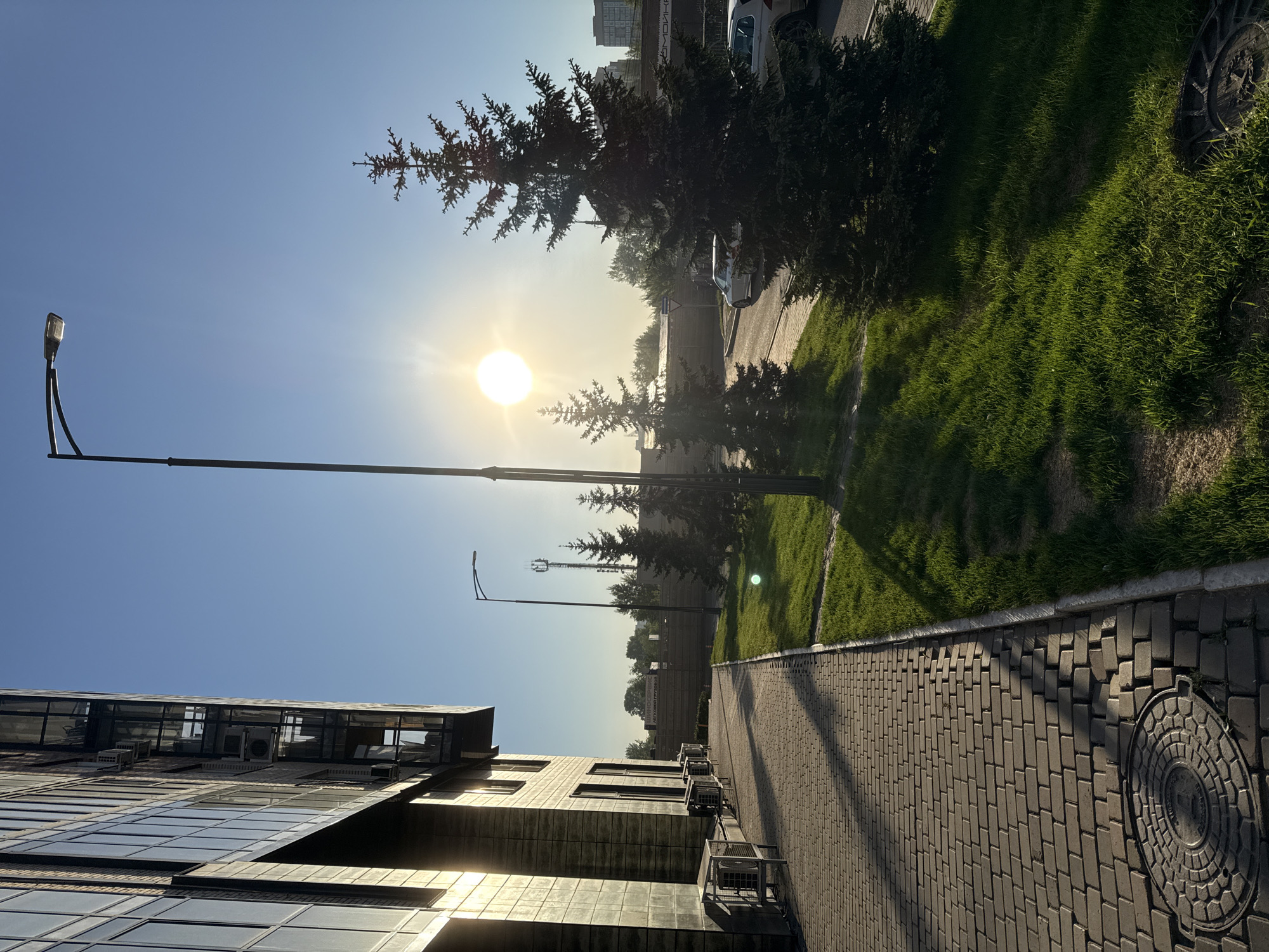 The image presents an urban or suburban street scene in Krasnoyarsk, Russia, captured under a clear, bright blue sky. The sun, low in the sky on the right, shines intensely, creating pronounced lens flares and casting long shadows from objects like trees and the street lamp across the pavement, suggesting either early morning or late afternoon.
In the foreground and extending towards the center, a neatly paved sidewalk made of grey rectangular bricks is visible. A circular metal manhole cover is embedded in the pavement in the immediate foreground. To the left of the sidewalk, a strip of vibrant green grass runs parallel, interspersed with several dark green evergreen trees and shrubs.
On the far left, a modern building features a facade of reflective glass windows and grey cladding, with multiple outdoor air conditioning units mounted on its exterior. A tall, black street lamp post with a modern light fixture stands prominently on the left side, its arm extending over the sidewalk. Another, smaller lamp post is visible further down the street.
Beyond the green space and trees, a road with several parked cars, including a white sedan, can be seen. In the distant background, urban structures and what appears to be a cellular tower are faintly visible. There are no people present, and no explicit activity is taking place, contributing to a calm and serene atmosphere. While some blurry text appears on a distant sign or billboard, it is indecipherable.