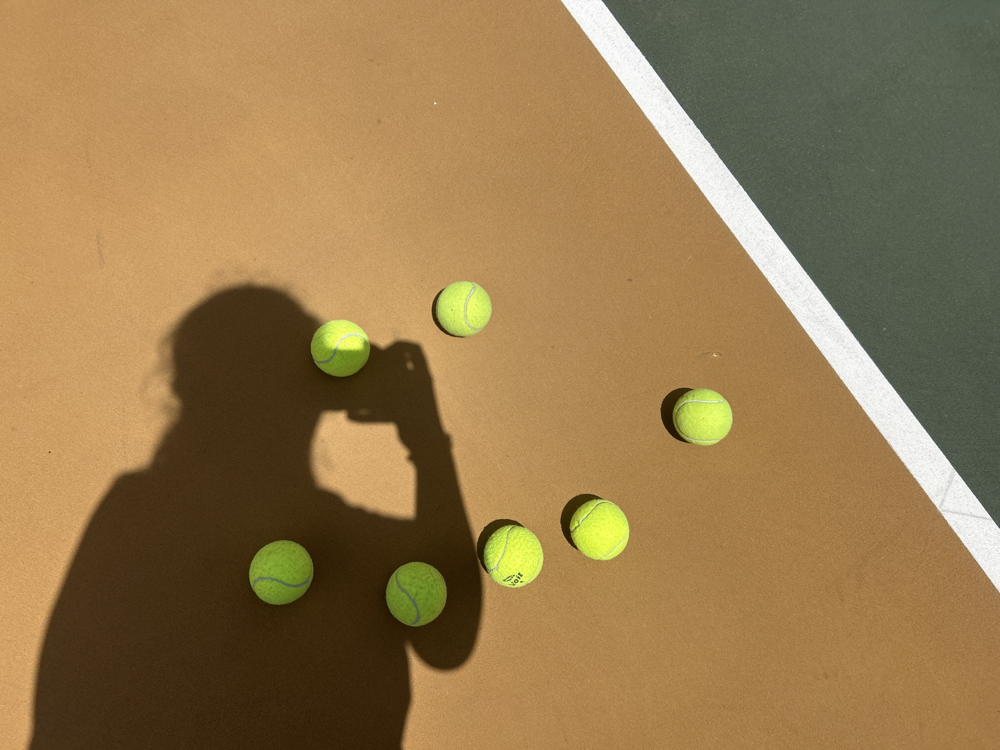 The image shows a section of a tennis court in the city of Puebla, Mexico, on a sunny day. The court is multi-colored, with a brown surface and a green section separated by a white line. Six tennis balls are scattered on the brown section. The shadow of a person holding a phone is cast on the brown surface, partially obscuring the tennis balls.