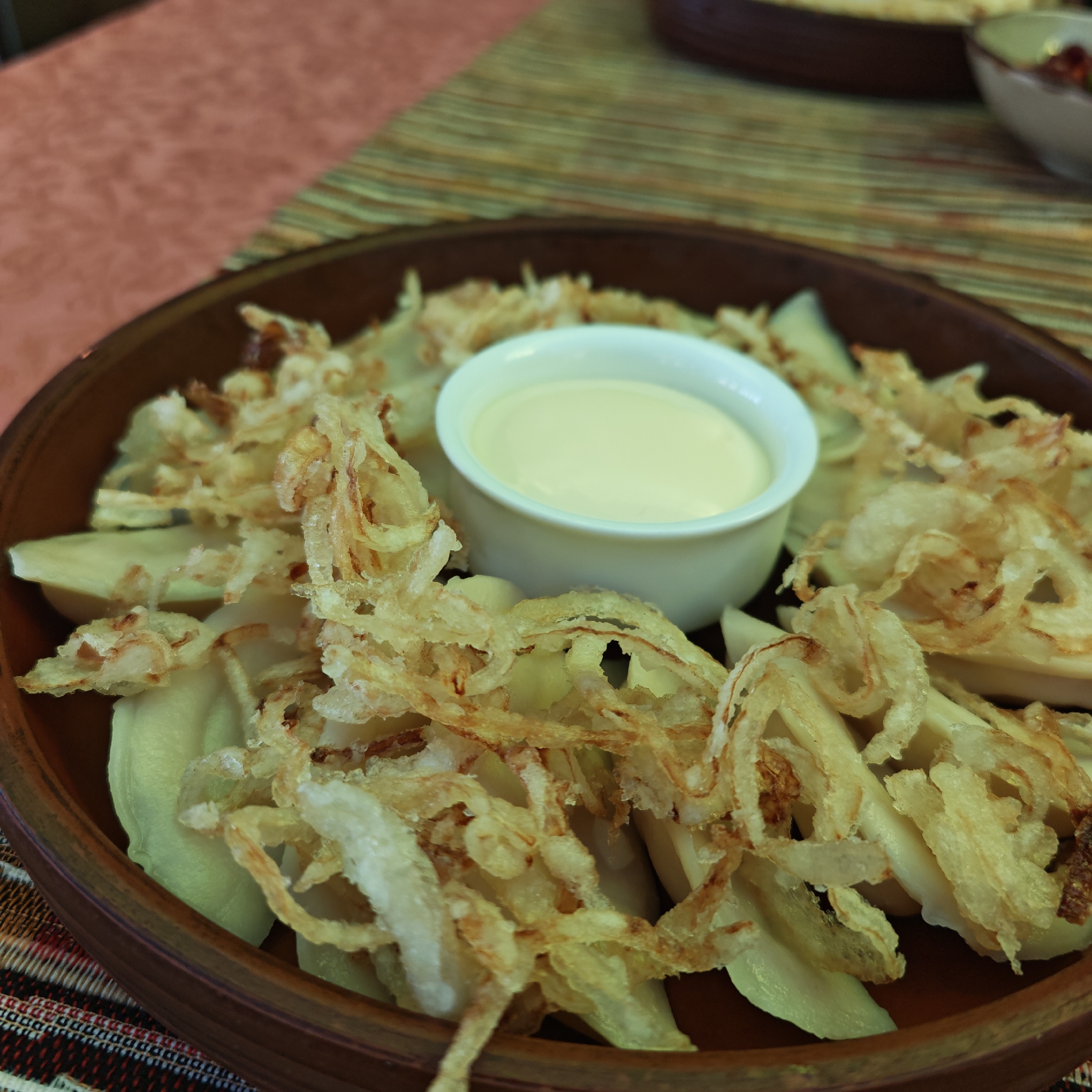 A close-up shot presents a wooden bowl filled with what appears to be dumplings or similar pasta, garnished with fried onion rings and accompanied by a small bowl of a creamy white sauce. The bowl sits on a multi-colored tablecloth with a striped pattern. In the background, partially visible, are elements suggesting a restaurant setting, possibly in Rostov, Russia. The lighting is diffused, highlighting the textures of the food and the surrounding materials.