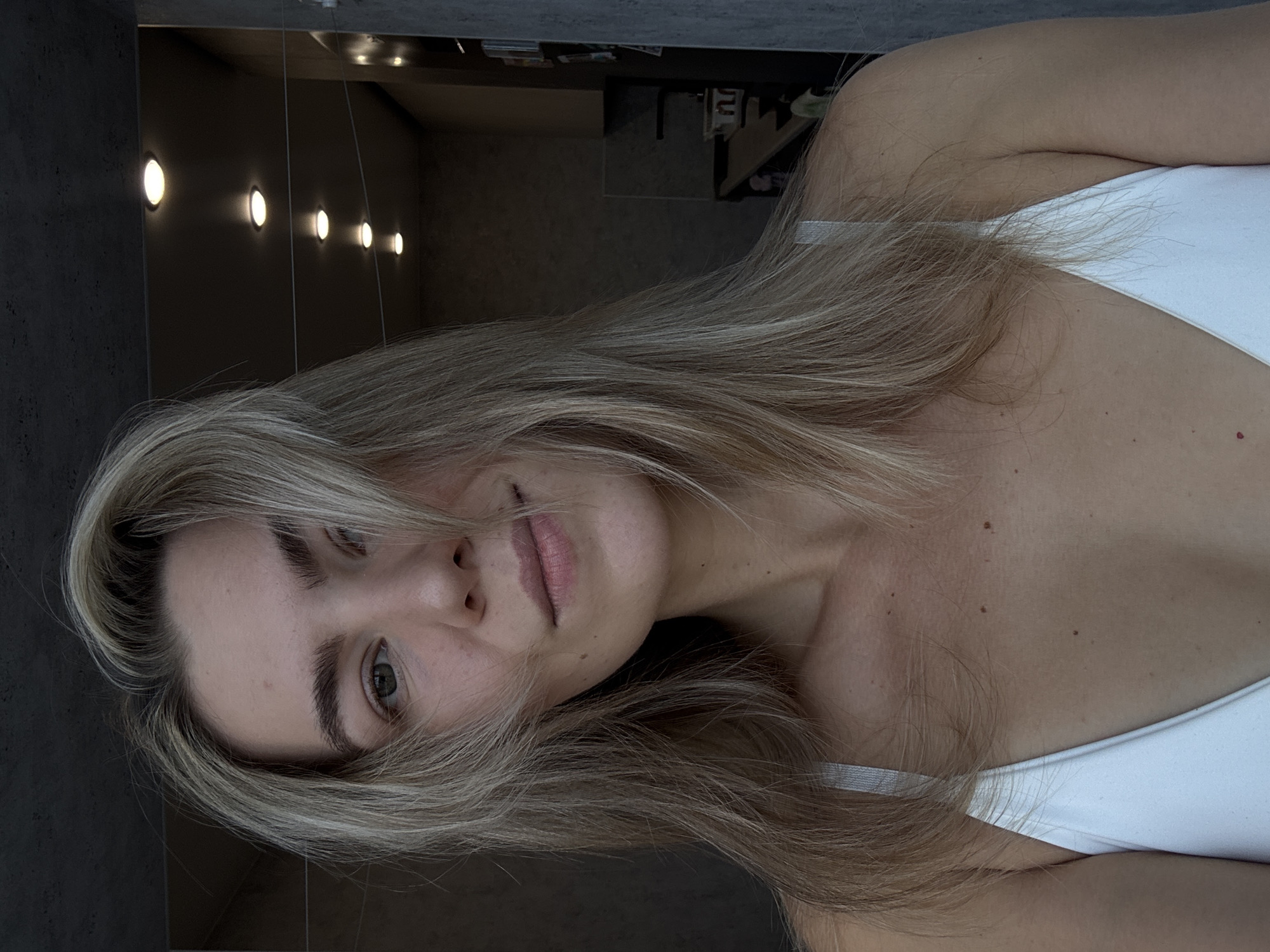 A woman with light blonde hair and a white tank top is the primary subject of the image. Her hair partially covers her face. The background includes a wall with several small, round lights. The location is a bathroom, evidenced by the vanity.