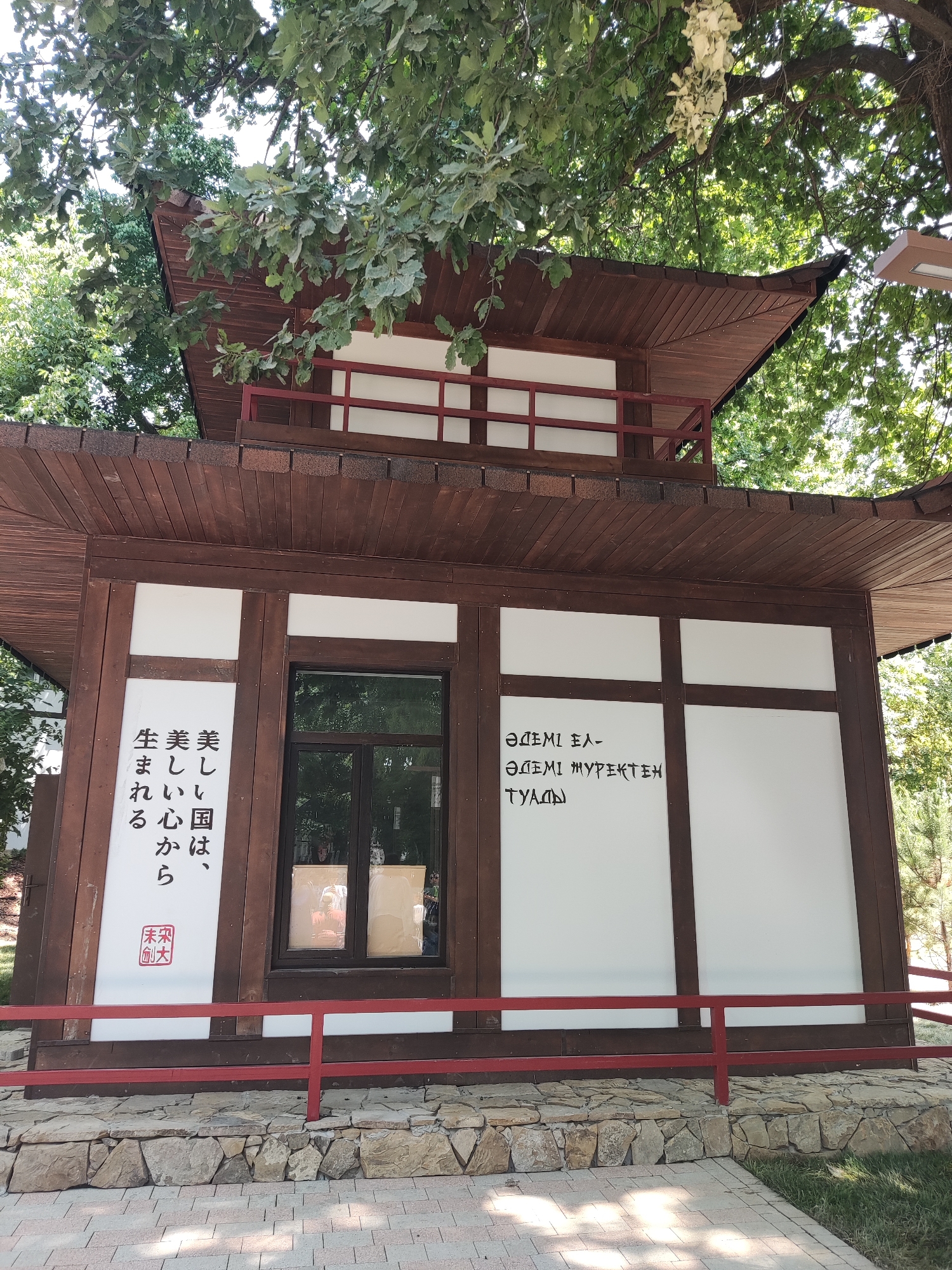 The image depicts a small, traditional-style Japanese building with a dark wooden exterior and a layered, dark brown roof. The building features white panels on its walls, some of which are adorned with text. On the left side, there are vertical lines of text in what appears to be Japanese script. Below this text, a red square stamp is visible. To the right of a window, there is more text, this time in Cyrillic script. The building has a red railing in front of it. The structure is situated outdoors, with lush green trees providing shade from above. The foundation of the building is made of large, irregular stones, and the ground in front is paved with light-colored bricks. The scene appears to be located in a park or garden setting. This building is part of the Japanese Garden in the city of Shymkent, Kazakhstan.