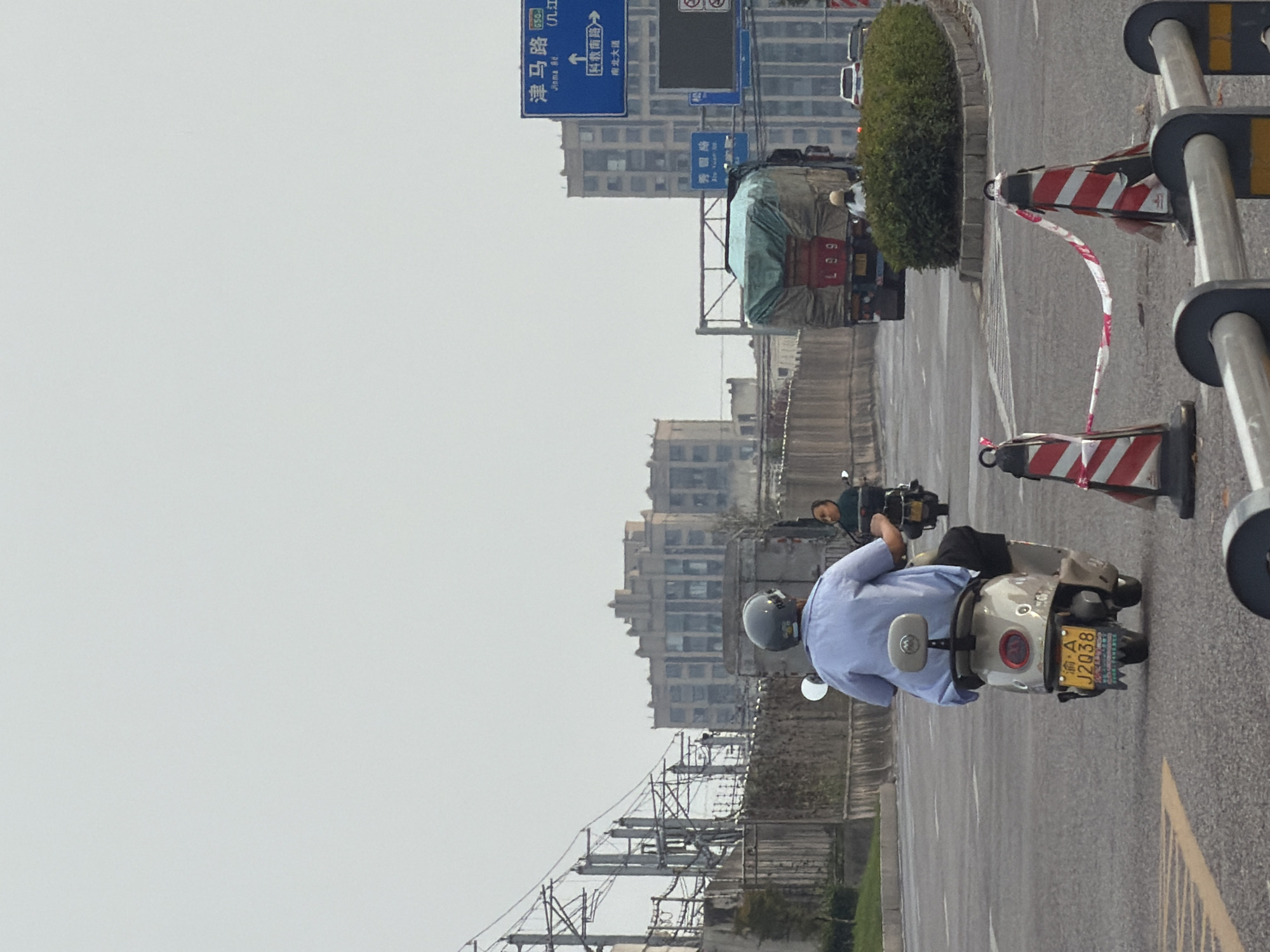 In Jiangjin, China, the image captures a hazy urban scene on an overcast day, likely during daylight hours. A person in a light blue shirt and a helmet is riding a beige scooter, distinguished by the license plate "渝A J2038" and a decorative tail. Following closely behind is another scooter carrying two individuals. In the background, a large truck covered with a green tarp, displaying "L 89" on its rear, proceeds down the road. A white car is visible further ahead, suggesting moderate traffic. Signage indicating directions to "Jinma Road" and "Kebei South Road" is present, along with directional arrows. The environment is characterized by asphalt roads, a concrete divider, and lush green bushes. Construction cones with red and white tape are placed on the side of the road, indicating a potential construction zone or road closure. Power lines and industrial-looking buildings are visible in the distance, contributing to the urban landscape.