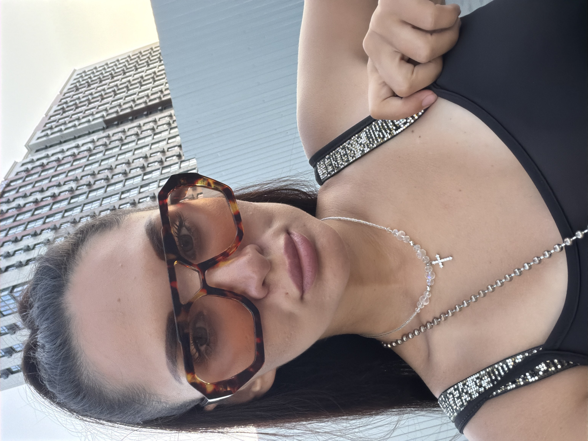 A woman is taking a selfie outdoors in front of a modern apartment building. She is wearing large, tortoise-shell framed sunglasses with amber lenses. She has dark hair pulled back, and is wearing a black tank top with sparkly straps and two necklaces, one of which has a small cross pendant. She is touching the strap of her tank top with her right hand. The background shows the side of a large, multi-story building with many windows and air conditioning units, and a pale blue fence or wall. The lighting suggests it is late afternoon or early evening. There is no indication of the location being the city of Azov, Russia.