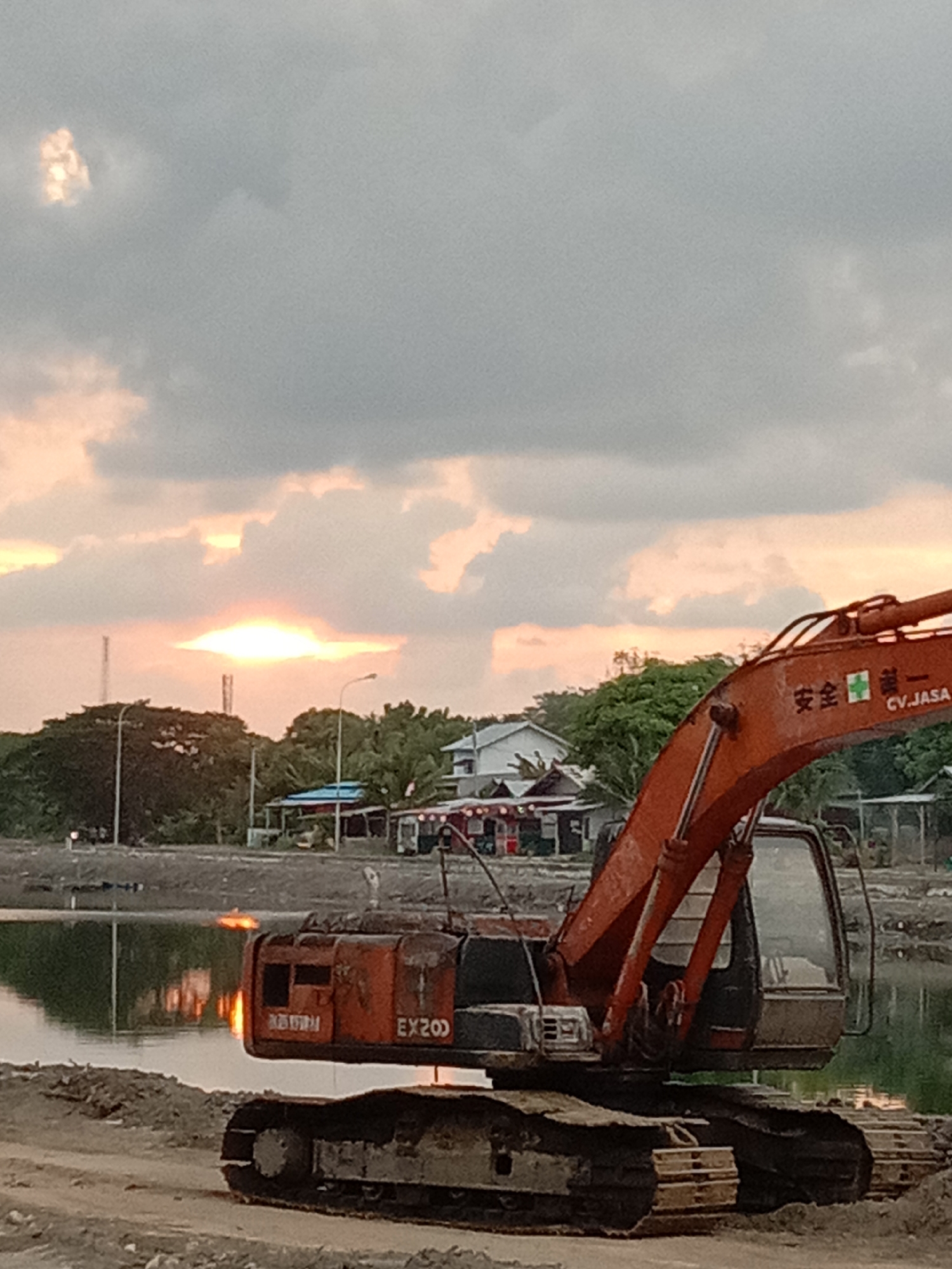 This is a photograph of an orange excavator positioned near a body of water, with buildings and trees in the background. The sky is overcast with a sliver of sunlight visible. The excavator has the designation "EX200" on its side and appears to be stationary. The scene is likely a construction site or an area undergoing development. The presence of a body of water suggests it could be near a river, lake, or coastal area. The buildings in the background appear to be of modest size, and some have red roofs. The overall impression is one of a developing landscape at dusk or dawn.