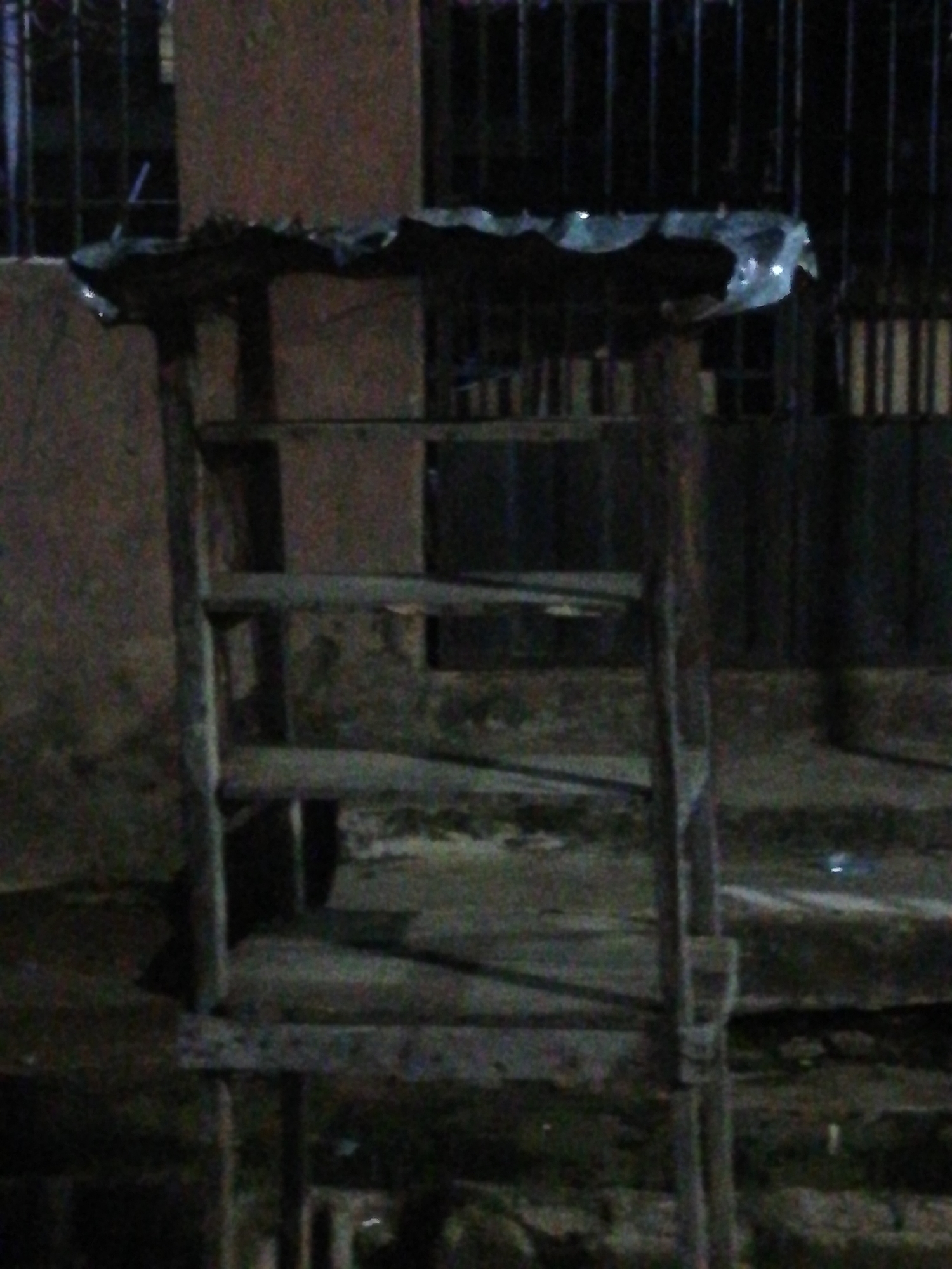 The image depicts a makeshift wooden structure, possibly a stand or display, positioned in an urban environment. It's comprised of vertical supports and horizontal shelves, with what appears to be a tattered covering on top. The structure exhibits signs of wear and age. The background features a gated metal fence. Given the information about location being Lagos, Nigeria, the setting suggests a street-side vendor setup. The lighting is dim and the scene has a gritty, urban feel.