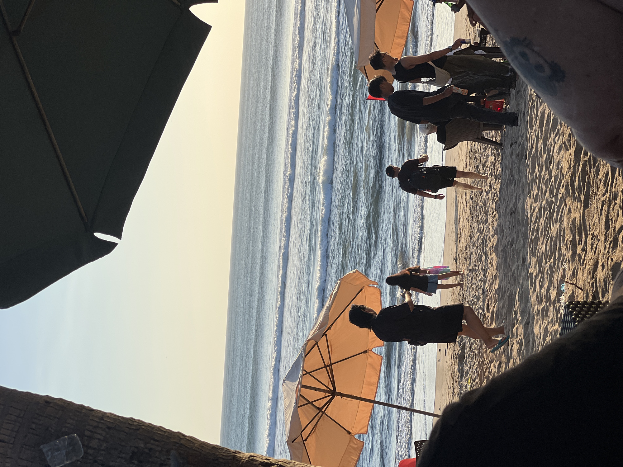In Kuta, Indonesia, at sunset, a beach scene unfolds under the soft golden light. The ocean stretches to the horizon, with gentle waves rolling onto the sandy shore. Several people are visible on the beach, some walking, others seated under umbrellas.
To the left, the rough texture of a palm tree trunk frames the view, with a small piece of plastic attached to it. In the foreground, a dark, out-of-focus object, possibly a piece of furniture or clothing, occupies the bottom left and right corners.
In the middle ground, an orange beach umbrella stands prominently, casting a shadow on the sand. A person in a black shirt and shorts stands near this umbrella, facing away from the camera and towards the ocean. Further back, a person in a dark shirt and shorts walks towards the water, and a person in a blue skirt and dark top walks away from the water.
To the right, another beach umbrella, partially visible and lighter in color, is present. Several individuals are gathered near it. Two men in dark clothing stand, one with a bag slung over his shoulder, and another in a black tank top and green pants also stands. A person is seated in a chair near this group.
The sand is a light brown, with footprints and scattered debris visible. The sky transitions from a pale yellow near the horizon to a soft blue overhead, indicating the late hour of the day.