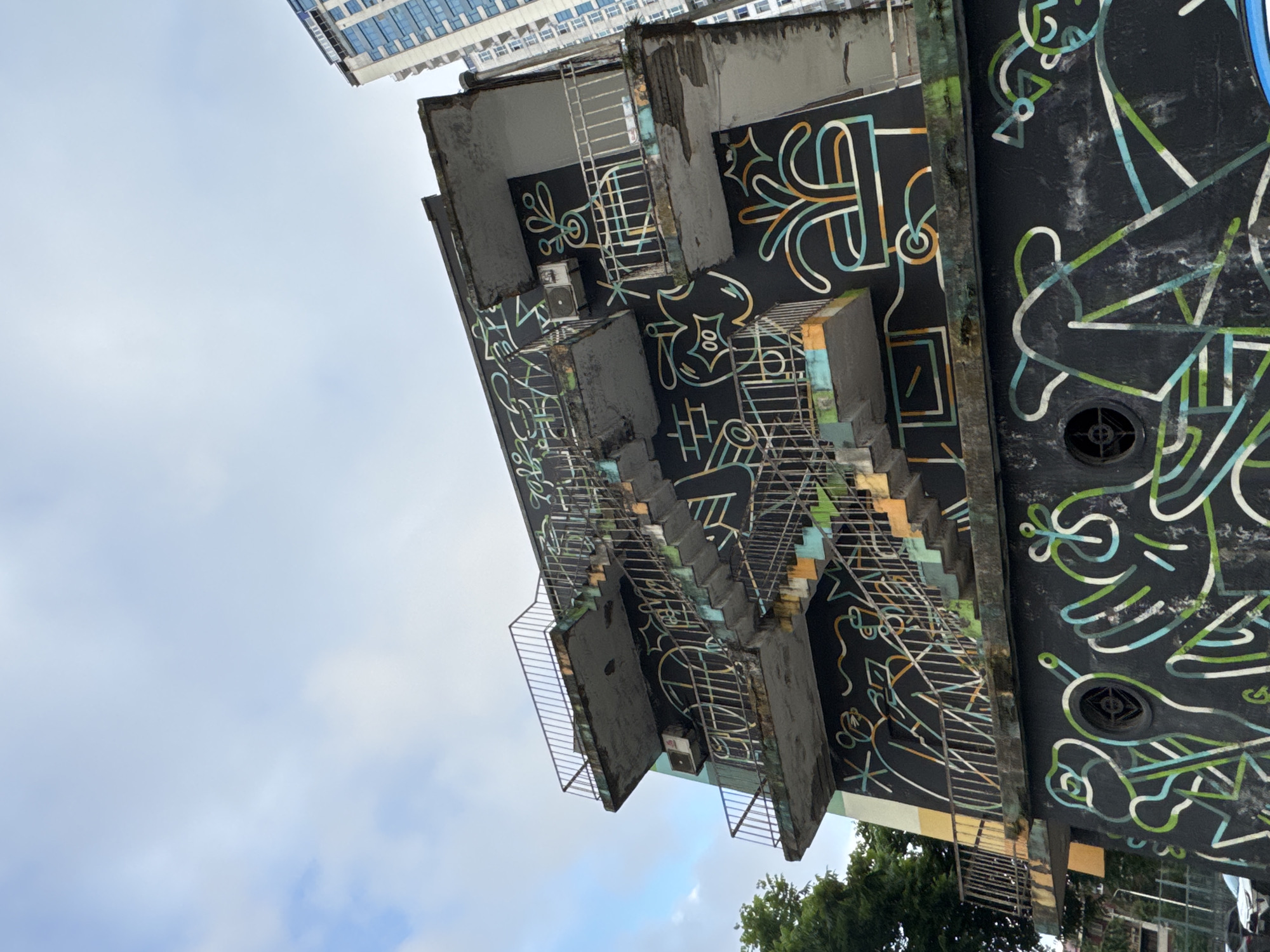 The image shows a concrete building with an external staircase, covered in black paint and adorned with colorful, abstract line art. The art features stylized figures, plants in pots, and geometric shapes in white, green, blue, and orange. The building appears weathered, with some peeling paint and rust on the metal railings of the balconies and stairs. An air conditioning unit is mounted on the wall of the second floor. In the background, a modern, tall building with blue-tinted windows is visible, suggesting an urban setting. The sky above is overcast with patches of blue and white clouds. This building is located in Batumi, Georgia, and is a notable example of street art in the city.