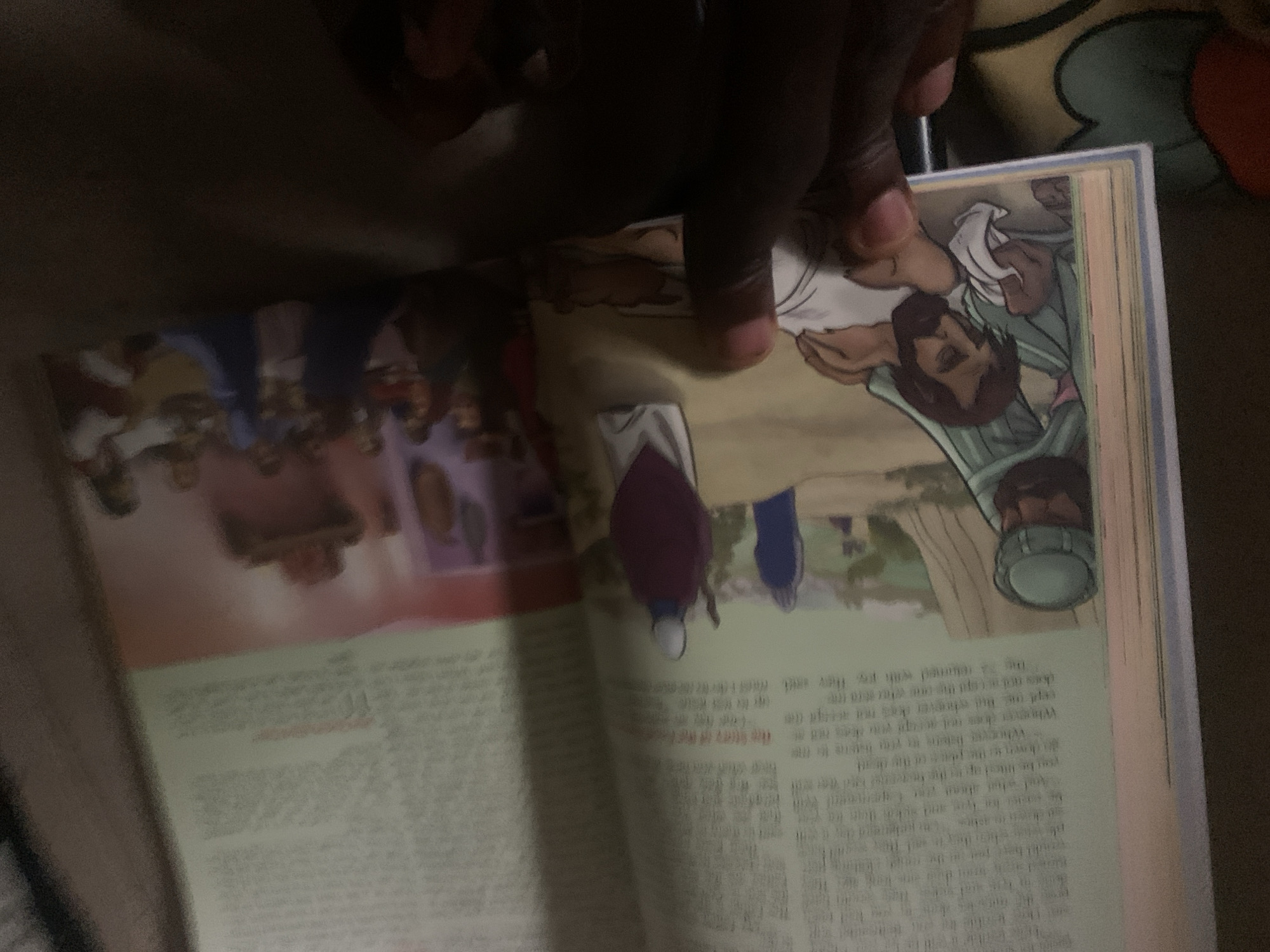 A close-up, angled shot captures an open book, with pages showing illustrations and text. The book is likely religious in nature, as evidenced by the depictions of figures that appear to be from biblical scenes, including a scene with Jesus healing a sick person. The lighting is dim, suggesting the photo was taken indoors or in low-light conditions. The book is lying on a patterned surface, possibly a blanket or rug. A person's hand is visible in the lower right corner, obscuring part of the book. The image does not provide any information about the city of Jauro Yinu, Nigeria.