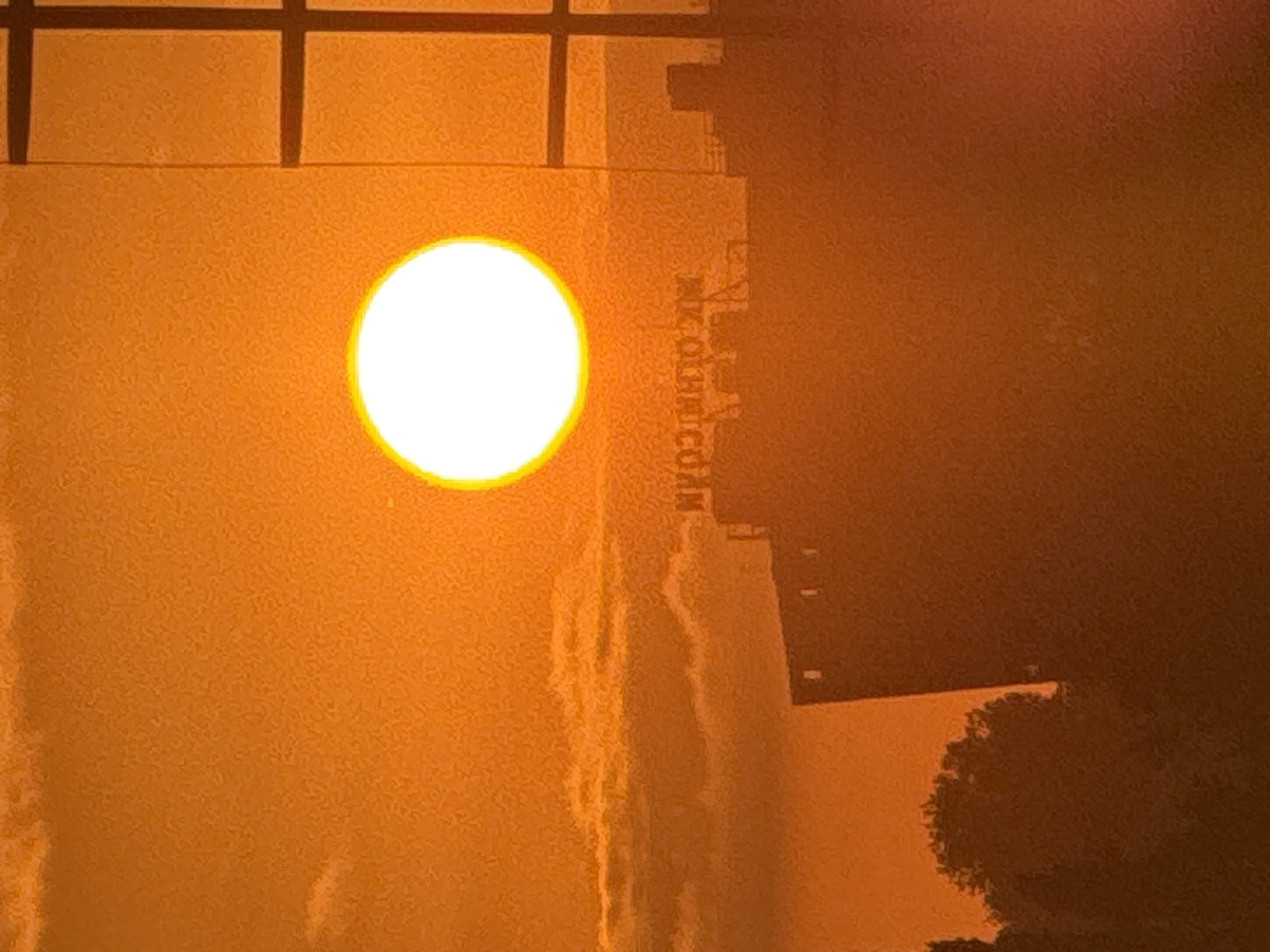This media file captures a sunset over a building in Sochi, Russia. The sun, a brilliant white disc with a warm orange halo, is prominently featured on the left side of the frame. The light from the sun casts an orange glow across the textured surface of the building, creating a warm and atmospheric scene.
The building appears to be a modern structure with a grid-like pattern of dark lines across its upper section, suggesting windows or architectural elements. On the right side of the building, there is some faint text visible, which is difficult to decipher due to the low resolution and the lighting conditions. It appears to be a vertical inscription.
The lower right portion of the image shows a dark silhouette of a tree or foliage, partially obscuring the building and the ground. The overall mood is serene and perhaps a bit melancholic, characteristic of a sunset. The warm colors suggest a clear or partly cloudy sky, typical of good weather. The scene is likely taking place in the late afternoon or early evening as the sun descends. The setting is urban, given the presence of the building, but with a touch of nature from the tree silhouette.