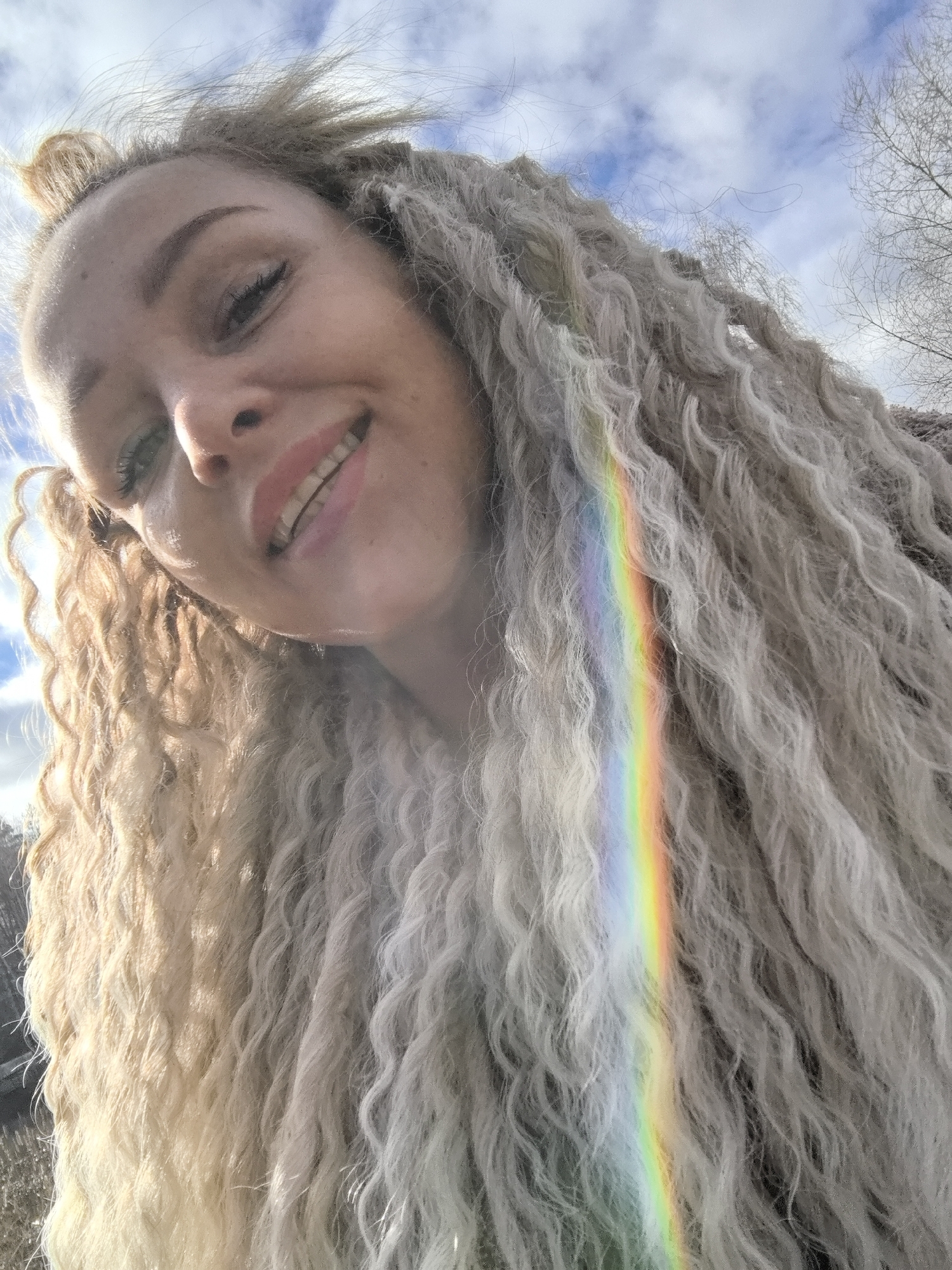 A selfie of a blonde woman with dreadlocks, smiling. A rainbow appears to be reflected onto her hair. The background shows a sky with clouds and some bare trees, suggesting an outdoor location in Noviy, Russia.