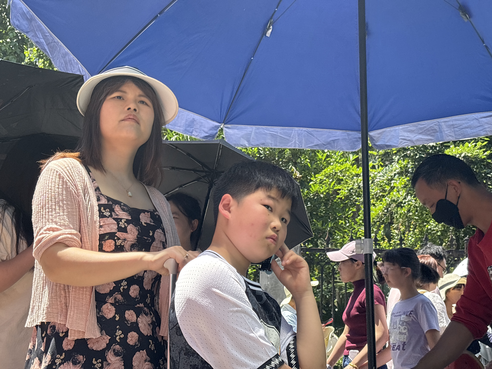 A crowded outdoor scene in Jiulongpo District, China, shows people sheltering under umbrellas. A woman in a floral dress, cardigan, and wide-brimmed hat stands next to a boy in a white shirt. The boy has his hand to his face in a contemplative gesture. Other people are visible, including a man wearing a face mask. Green foliage is in the background. The sky is bright, suggesting daylight hours.