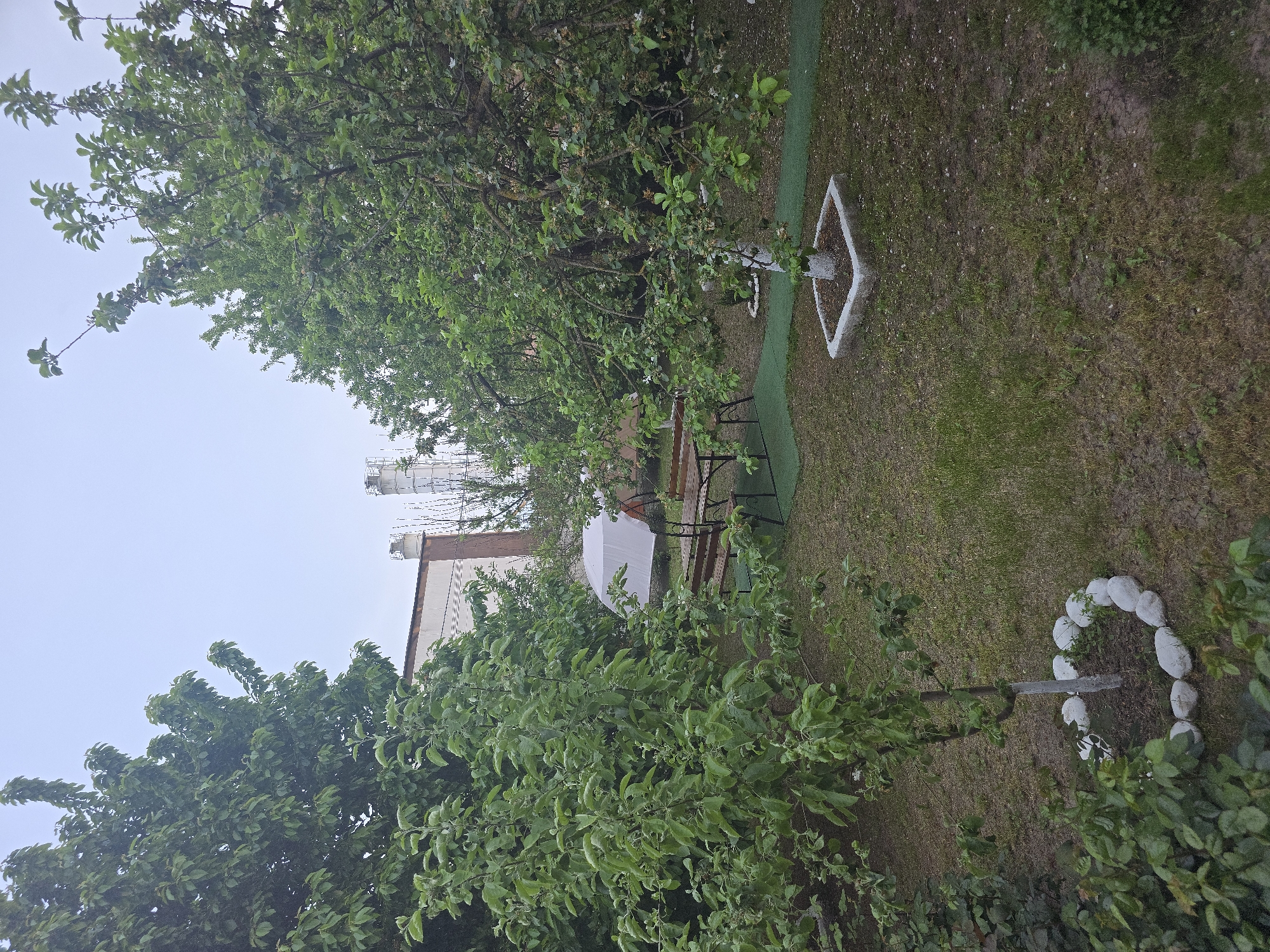 A medium-range, eye-level shot depicts a garden. The garden features a patch of grass that appears uneven in color, alternating between green and brown, suggesting varied sunlight exposure or differing grass types. A table and several chairs are set up in the middle of the garden. The table is covered with a white canopy. Surrounding the garden are various trees and plants, with some bearing fresh green leaves. There are white stones arranged in a circle. There's a building with tanks visible above it. A diamond-shaped concrete structure sits near a tree in the garden. The scene is set in the city of Unknown, Unknown.