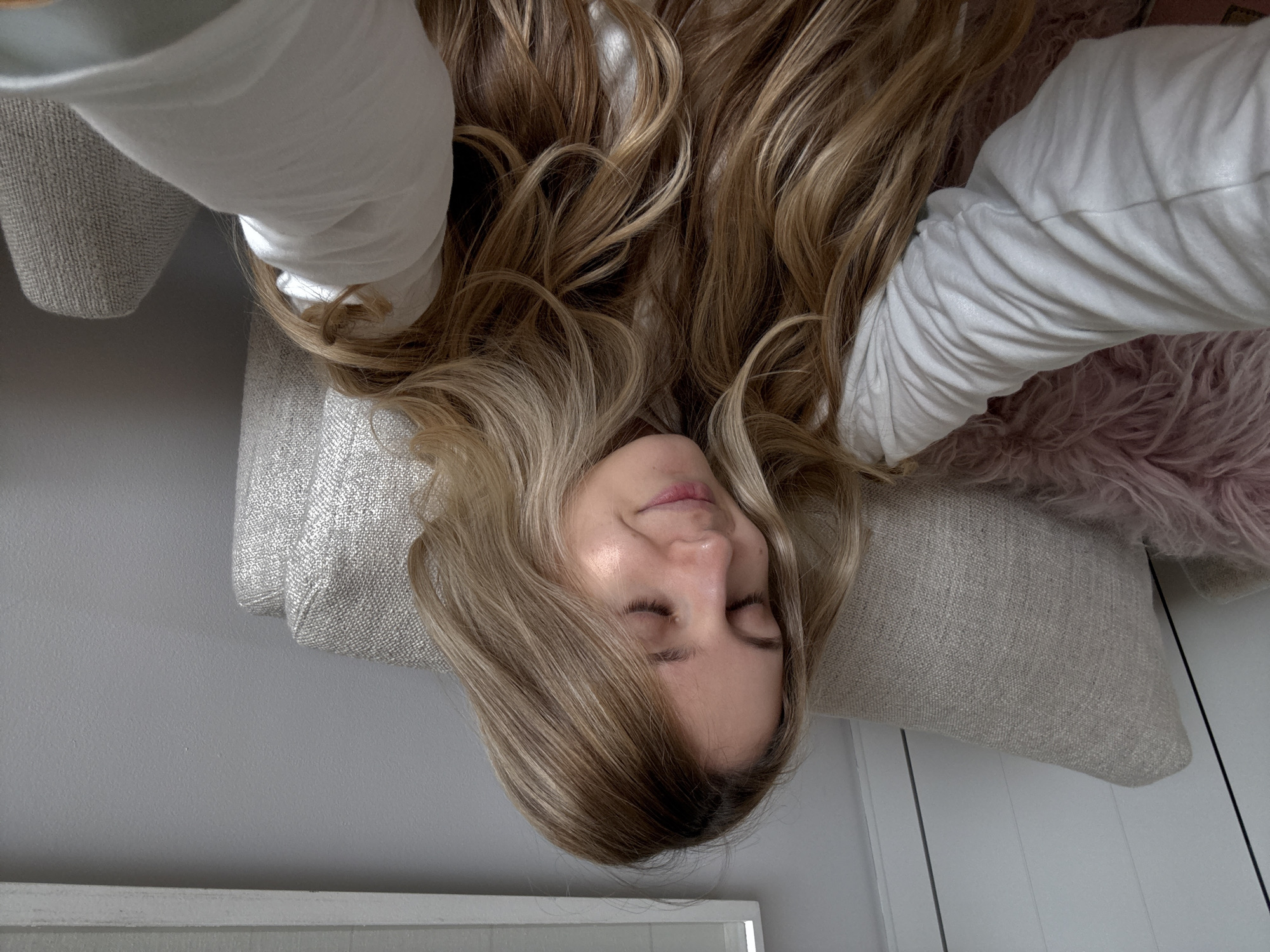The image shows a woman resting indoors, presented in an inverted perspective where her head is positioned towards the bottom of the frame. The woman has long, wavy blonde and light brown hair that cascades around her head and shoulders. Her eyes are gently closed, and she exhibits a serene, relaxed expression, suggesting she is either sleeping or in a state of deep rest. She is wearing a light-colored, possibly white or off-white, long-sleeved top.
Her head rests on a textured, light beige pillow or cushion. Another similar cushion is visible beneath her body on the left. To the right of her, a fluffy, pinkish or purplish textile, likely a throw blanket or decorative pillow, adds a soft element to the composition. The background features a plain light gray or off-white wall. At the very bottom of the frame, a white baseboard or furniture trim is visible. The lighting is soft and ambient, characteristic of an indoor setting with indirect light, making the precise time of day difficult to determine. There is no visible text in the image. The overall scene conveys a sense of peace and comfort. The location context provided for this image is Mexico City, Mexico.