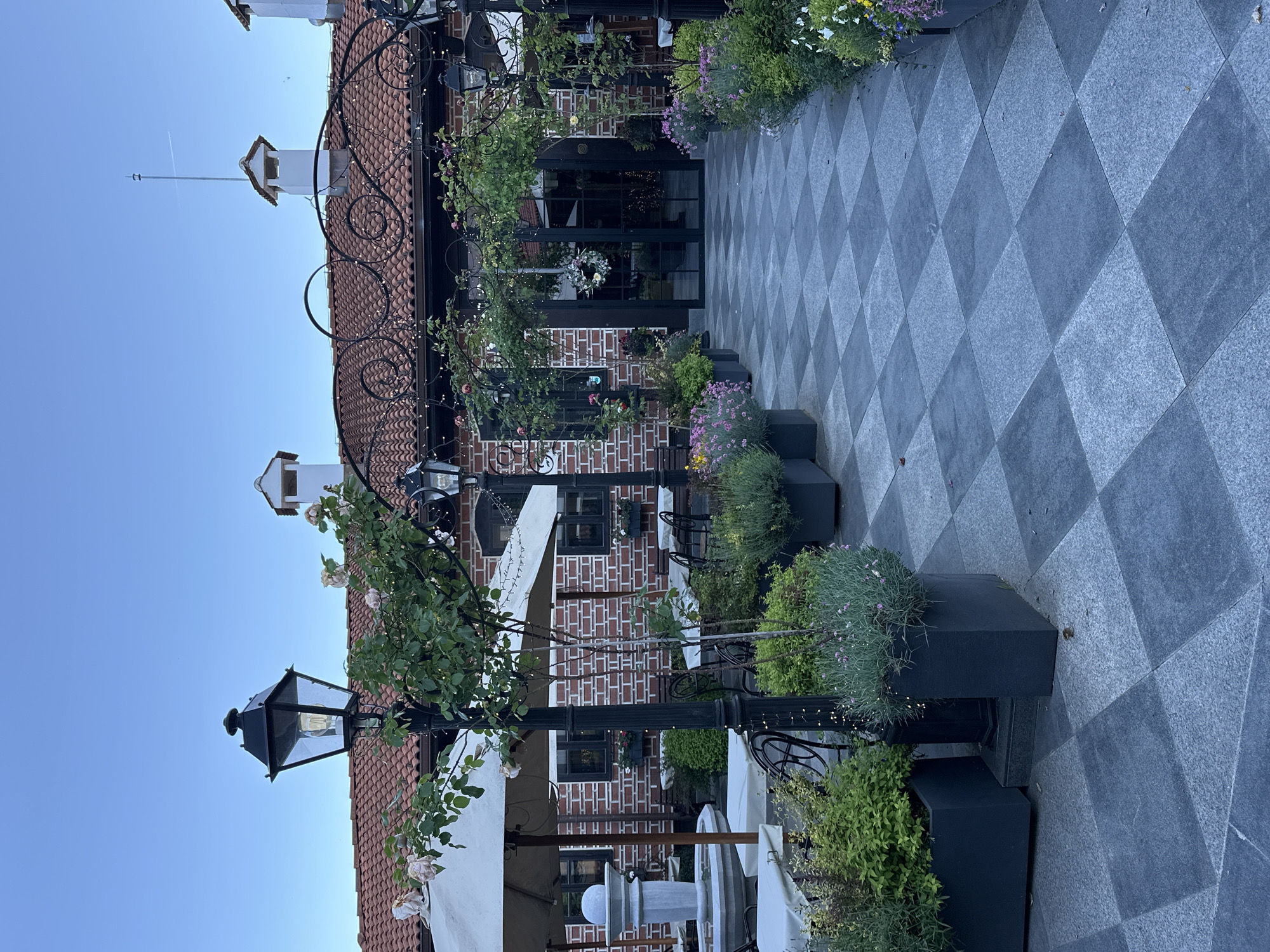 The image depicts an outdoor scene at a restaurant in Veliko Tarnovo, Bulgaria. The building is brick with a red tile roof and white trim. The ground is paved with gray and dark gray diamond patterned stones. There are black planters lining the walkway, containing green plants and purple flowers. Wrought iron arches adorned with greenery and lights span the walkway leading to the restaurant entrance. Tables and chairs are visible under white umbrellas. A lamp post stands at the left side of the image. The sky is clear and blue.