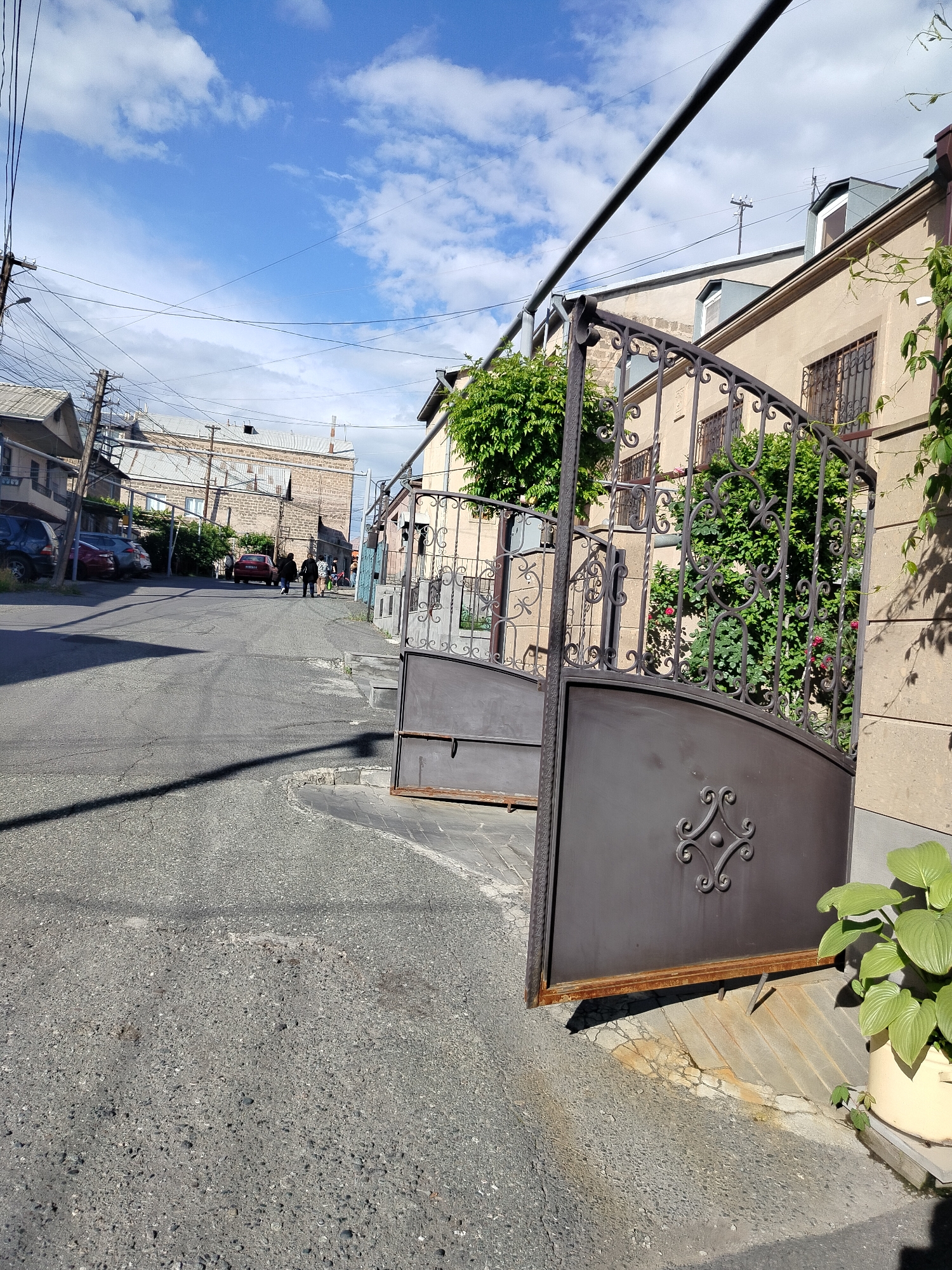 An eye-level shot shows a street in Yerevan, Armenia with an open wrought-iron gate in the foreground. The gate is attached to a beige building with barred windows and climbing greenery. A large plant in a pot sits to the right of the gate. The street is asphalt and slightly rough.
In the background, several cars are parked along the left side of the street. Two people are walking away from the camera down the street. Buildings are visible on both sides of the street. A network of electrical wires stretches across the sky. The sky is blue with scattered clouds.