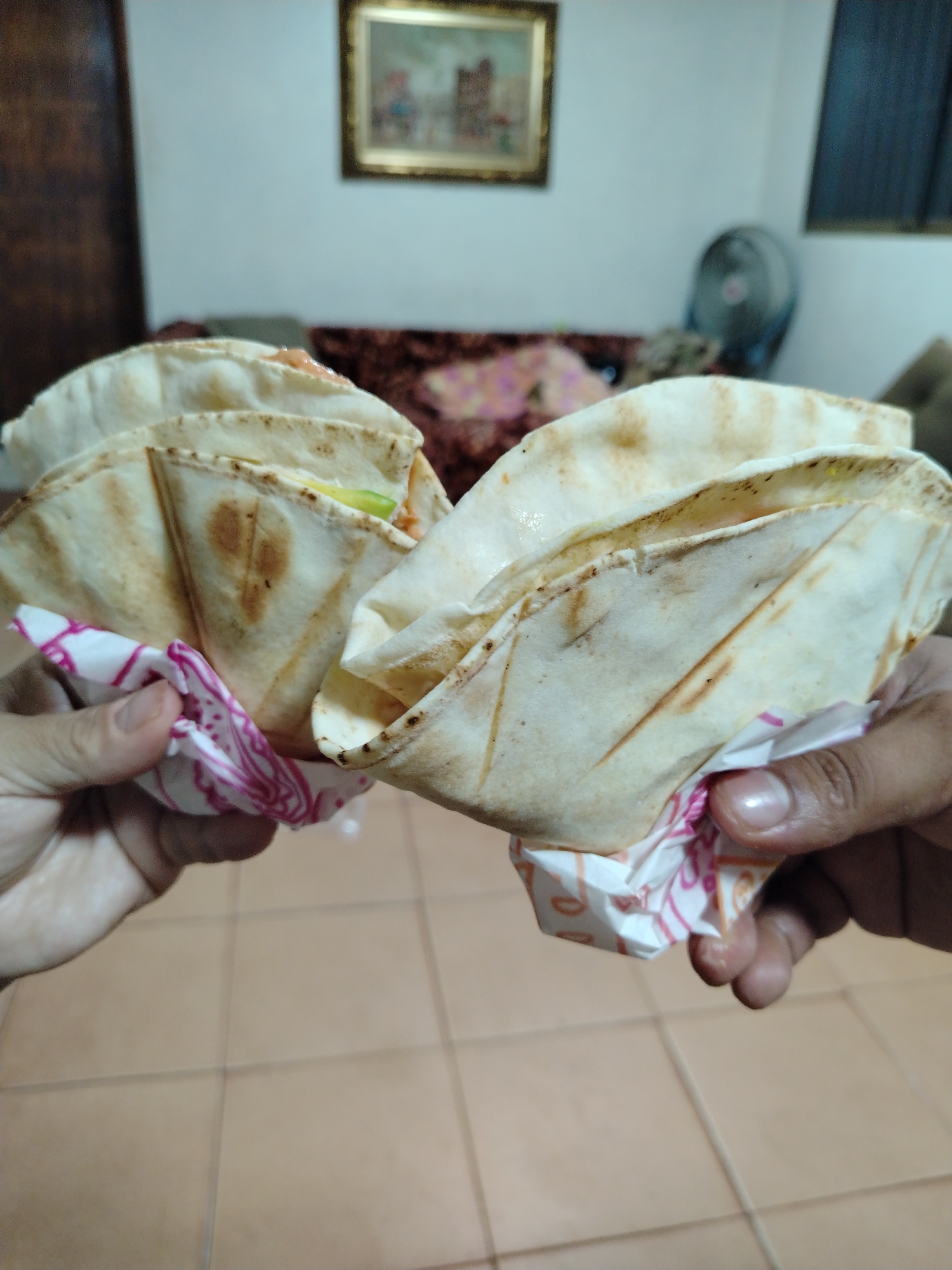 Two hands are holding filled pita bread wraps, each in a white paper wrap with pink designs. The background includes a tiled floor, a couch, a painting on the wall, and a fan. The image was taken indoors in Tinaquillo, Venezuela.
