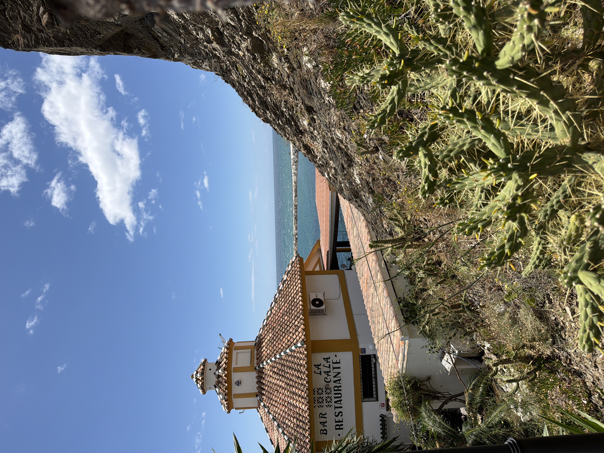 The image presents a view of the "Bar La Cala Restaurante," located in Benalmádena, Spain, as seen through a natural arch or opening in a stone structure. The restaurant building has a white facade with a tiled roof in shades of red and orange, and the words "BAR LA CALA RESTAURANTE" are visible on the side of the building. The scene includes a view of the sea and the sky, partly obstructed by the opening. Cacti and other vegetation are visible along the edges of the opening. No people are visible in the shot.