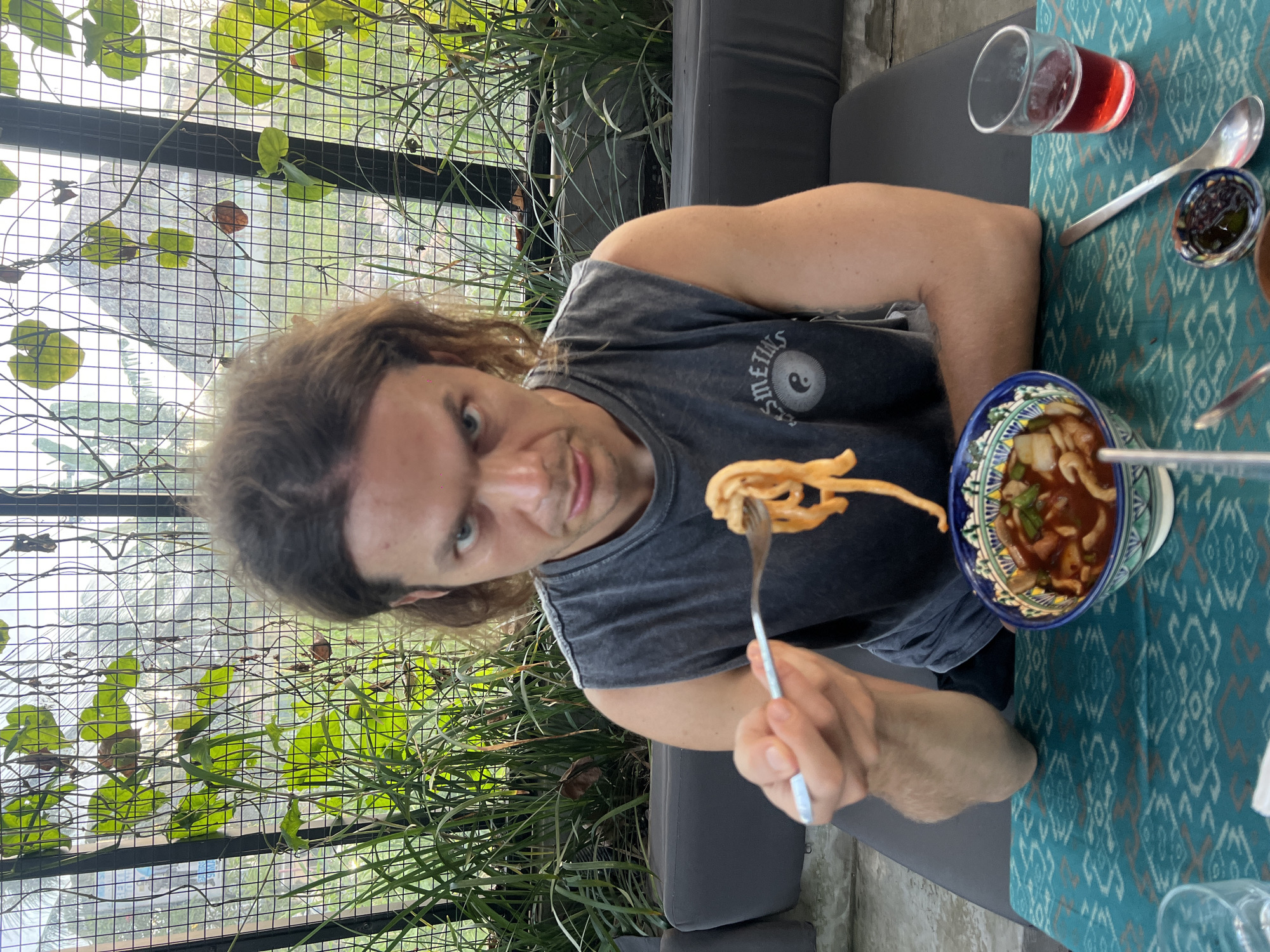A candid, medium-shot photograph shows a man dining in what appears to be an outdoor or semi-outdoor restaurant setting. The image is oriented vertically (rotated 90 degrees counter-clockwise from the original).
The primary subject is a man with long, wavy brown hair and light blue eyes, looking directly at the camera with a slight, playful smirk. He is wearing a dark grey, sleeveless t-shirt with a white circular logo on the left chest area, featuring a Yin and Yang symbol and partially visible text that reads "SMELLS". His right arm is bent, holding a fork with a mouthful of light-colored noodles or pasta, poised as if about to eat.
In front of him, on a table covered with a teal and white patterned tablecloth, sits a decorative bowl with intricate blue and white designs. The bowl contains a reddish soup or stew with various ingredients, including what look like sliced mushrooms and green herbs. To the right of the bowl, there's a clear glass filled with a red beverage, a silver spoon, and a small dish holding a dark condiment.
The background reveals a lush, green environment. Behind the man, a metal grid structure (possibly a trellis or fence) is covered with climbing green vines and leaves, suggesting an open-air or greenhouse-like dining area. Dark grey upholstered bench seating is visible on either side of the man, extending into the background.
The scene is brightly lit by natural light, indicating daytime. The general ambiance, with the vibrant greenery and casual dining setup, is consistent with the provided location context of North Kuta, Indonesia. The man's direct gaze and expression suggest an interaction with the photographer, possibly a friend or companion.