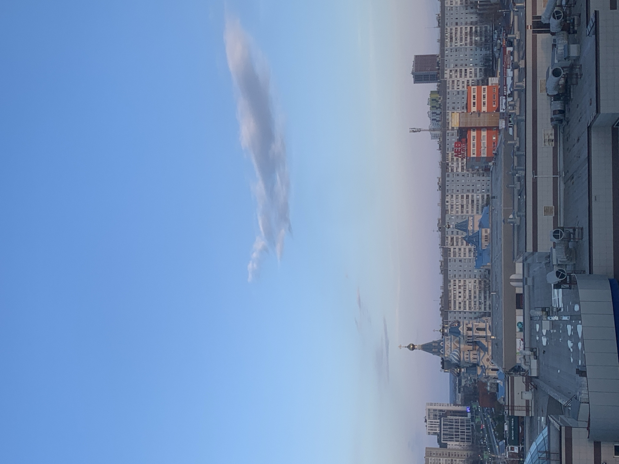 The image shows an elevated cityscape, likely taken from a high building in Izhevsk, Russia. The sky, occupying the left side of the image, is a clear light blue with a thin, vertical cloud formation. The cityscape consists of various buildings, including residential blocks in gray, white, and orange, as well as a prominent Orthodox church with multiple blue domes topped with gold crosses. The church is positioned slightly left of the center of the image. In the foreground, there is an industrial rooftop with vents and ducts. The cityscape fades into a hazy horizon, suggesting the photo was taken during the late afternoon or early evening.