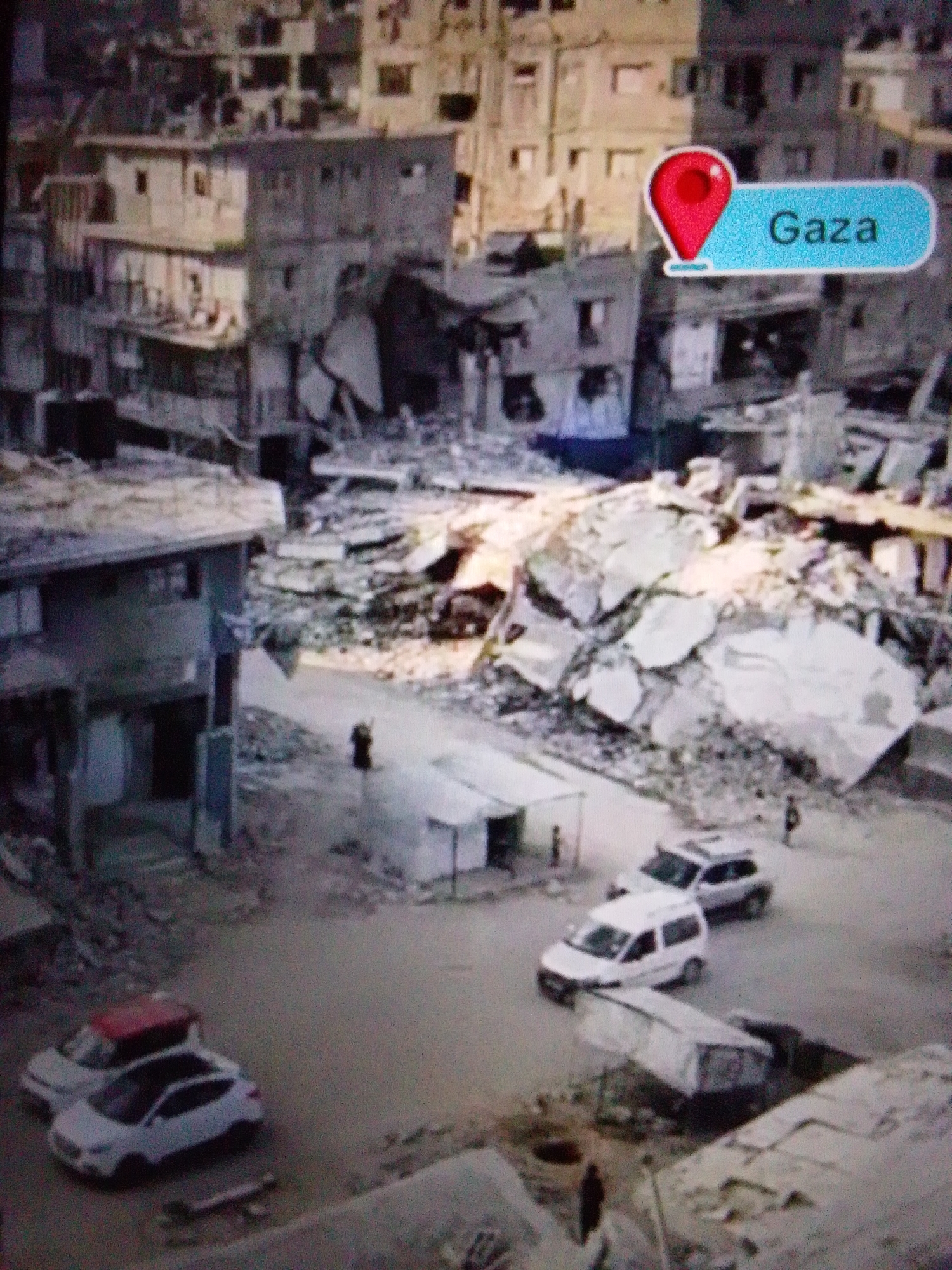 The image depicts a scene of widespread destruction in a city. Buildings are heavily damaged, with debris strewn across the ground. A map marker with the text "Gaza" is superimposed on the image, indicating the location. Several vehicles, including cars and what appears to be a van, are visible on a paved road amidst the rubble. A few individuals are seen walking in the vicinity. The overall environment suggests a recent catastrophic event. The city of Aznakayevo, Russia, is not depicted in this image.