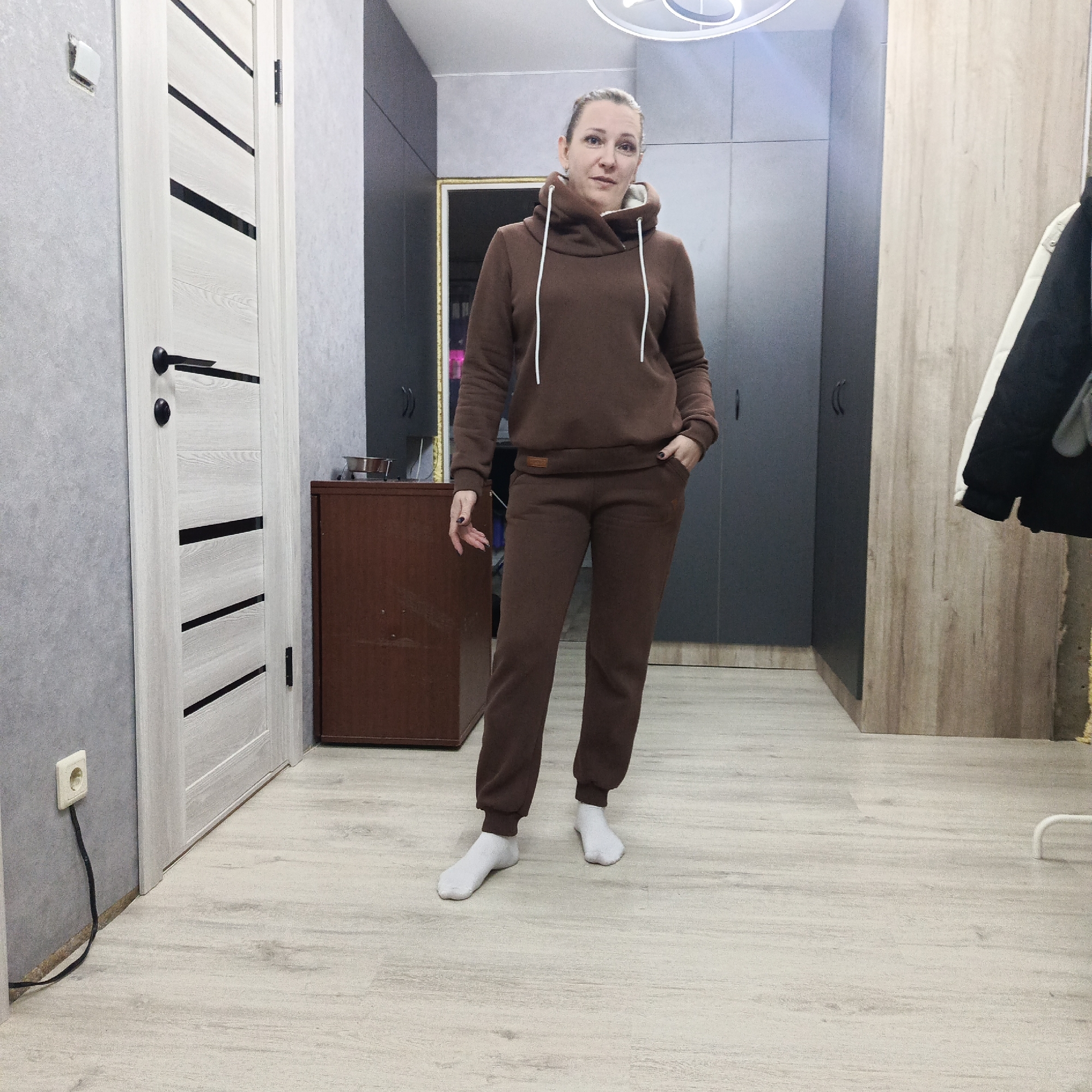 A woman stands posing casually in an indoor hallway or room in Markovskiy, Russia. She is the central subject, wearing a matching dark brown (chocolate brown) hooded tracksuit with prominent white drawstrings around the neck. Her left hand is tucked into her pants pocket, while her right hand hangs by her side. Her hair is pulled back, and she wears plain white socks, looking directly at the camera with a neutral expression.
The setting features a mix of modern interior design elements. The wall on the left is covered with light gray textured wallpaper. A contemporary white interior door with several black horizontal and diagonal stripes and black handles is visible on the left side of the frame. Near the bottom left, an off-white electrical outlet with a black cable plugged into it and extending downwards is present. The floor is light-colored wooden laminate.
To the right of the door, a dark brown wooden dresser or low cabinet holds a shiny metallic bowl on its top surface. Behind the woman, dark gray or charcoal-colored built-in cabinets or wardrobes line the wall. Further to the right, another section of light wood-paneled wall or cabinet is visible, with a black and white jacket or coat hanging on a hanger. A modern, circular white light fixture is affixed to the ceiling. A small, light brown brand label, likely made of faux leather, is sewn onto the lower front band of her sweatshirt, but no text is clearly legible. The scene is illuminated by artificial indoor lighting.