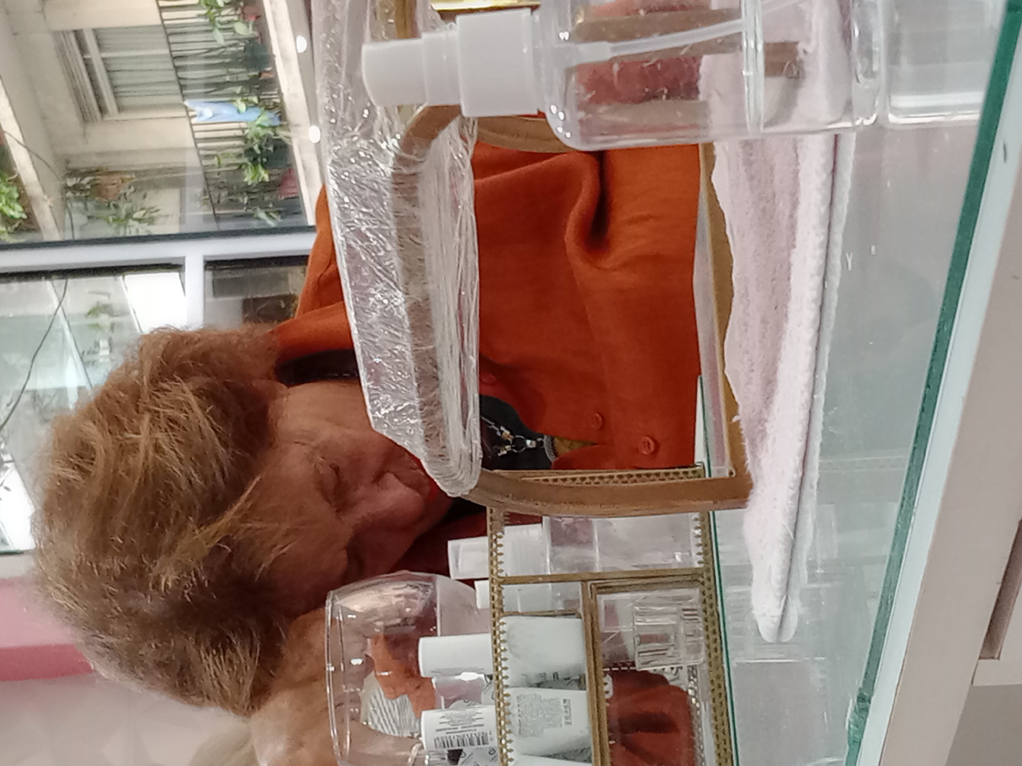 A mature woman with light brown, curly hair, wearing an orange top, is seen reflected in a mirrored vanity table. The table holds several clear plastic bottles and containers. The reflection also reveals a window with outdoor foliage, suggesting a location with natural light and possibly an indoor garden or courtyard. The setting could be a beauty salon or a personal vanity space. The location appears to be in Buenos Aires, Argentina, based on the general architectural style and available light typical of the city.