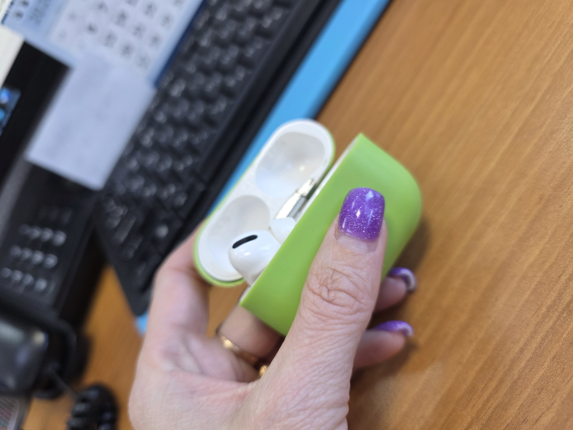 A hand is holding open a white AirPods Pro case covered with a green silicone sleeve. The AirPods Pro are visible inside the case. The hand has purple glittery nail polish. A black computer keyboard is visible in the background, resting on a blue surface. The photo is taken on a wooden surface, possibly a desk. Based on previous conversation history, the location may be in the city of Chaikovsky, Russia.