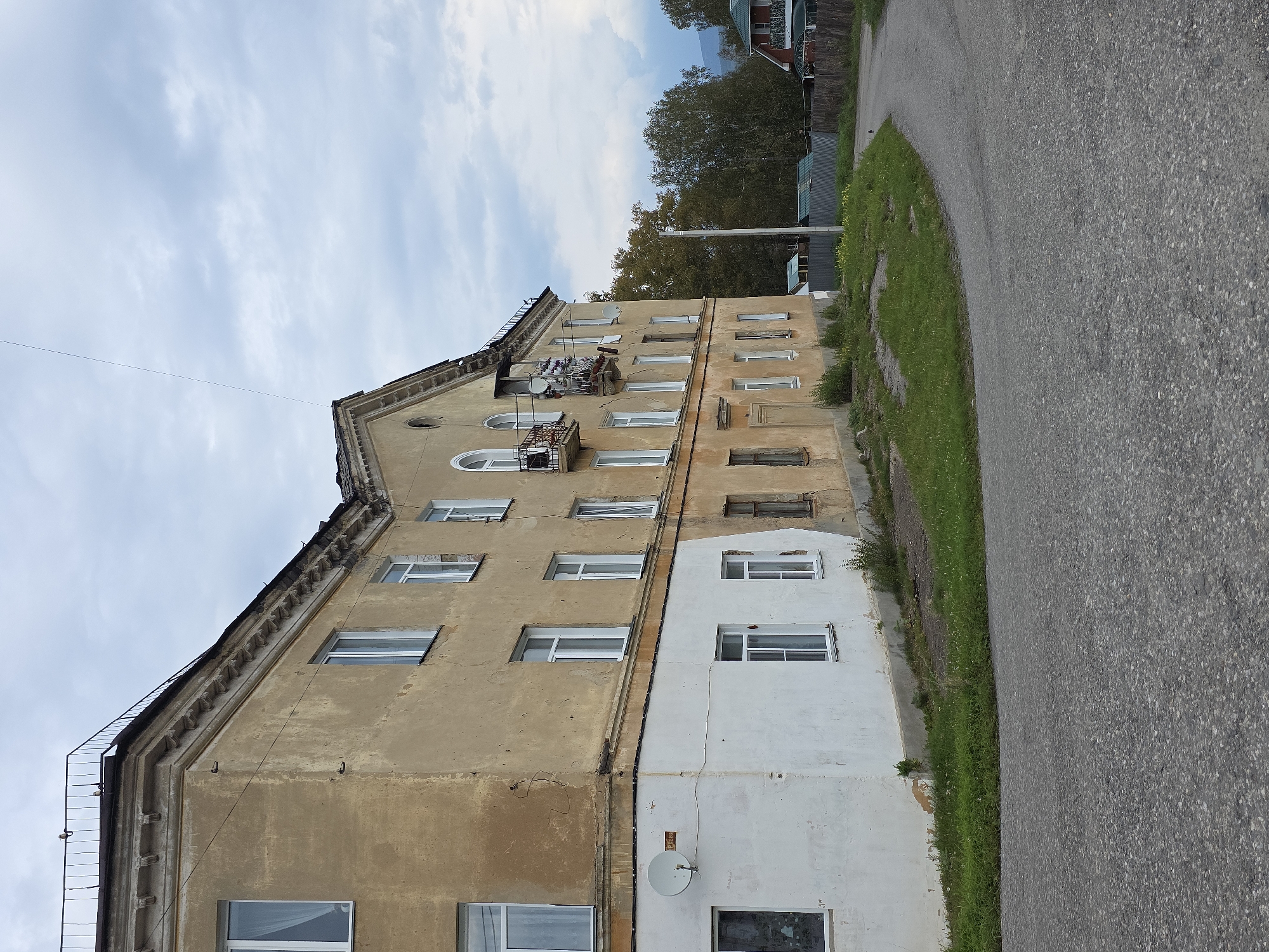 The image captures a view of a dilapidated, multi-story apartment building in Magnitka, Russia, under a cloudy sky. The building, painted in a faded yellow and white, shows signs of neglect with peeling paint and weathered facades. It features numerous windows, some with white frames, and balconies with rusty railings. A satellite dish is mounted on the lower white section of the building.
The building is situated on a slight incline, with a patch of overgrown grass and weeds in front of it. A paved road curves away from the building into the distance, disappearing behind a line of trees. In the background, other residential structures, possibly houses with red roofs, and a metal fence are visible, suggesting a semi-rural or suburban setting. The overall atmosphere is one of quiet decay and a subdued, overcast day. No people are visible in the scene.