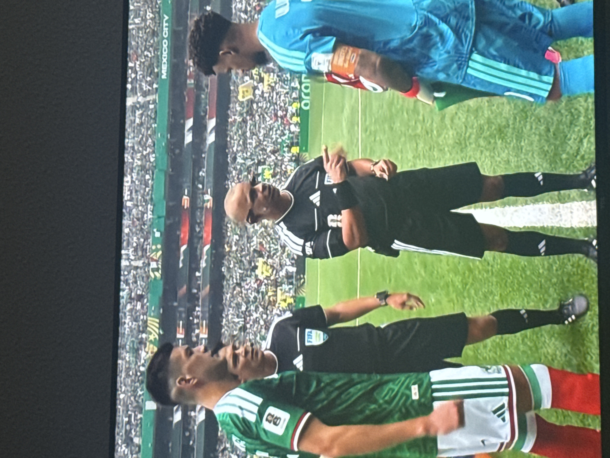 In an outdoor soccer stadium, three individuals are in the foreground on a grass field marked with white lines. A soccer player in a green and white striped jersey and red socks stands on the left. To his right, a referee in a black uniform with white stripes on the sleeves and shorts is speaking to the player. The referee is wearing a black watch and has a bald head. Another individual, presumably a player in a blue jersey and shorts, stands to the right of the referee, facing him. The background shows a dense crowd in the stadium seating, with some banners and signs visible, including one that reads "MEXICO CITY". The overall lighting suggests it is daytime.