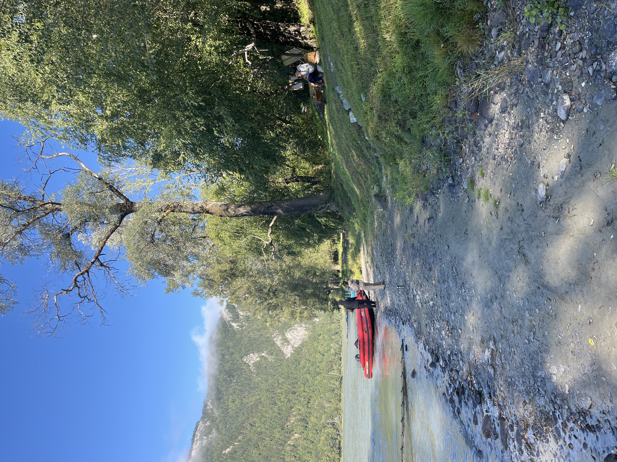A bright, sunny day at the shore of a wide, calm river in Ust-Muny, Russia. The water is a pale, murky green, with a rocky, sandy embankment. A bright red inflatable boat is partially submerged on the bank, with two individuals standing near it. One, wearing a black hooded jacket and dark pants, stands facing the boat. The other, in beige pants and a light-colored top, faces away from the boat, towards the water. Further inland, on a grassy slope shaded by mature trees, two more individuals are seated on a wooden bench, appearing to be engaged in conversation. The background features a lush green forest leading up to a mist-shrouded mountain range. The overall scene evokes a sense of peaceful recreation in a natural setting.
