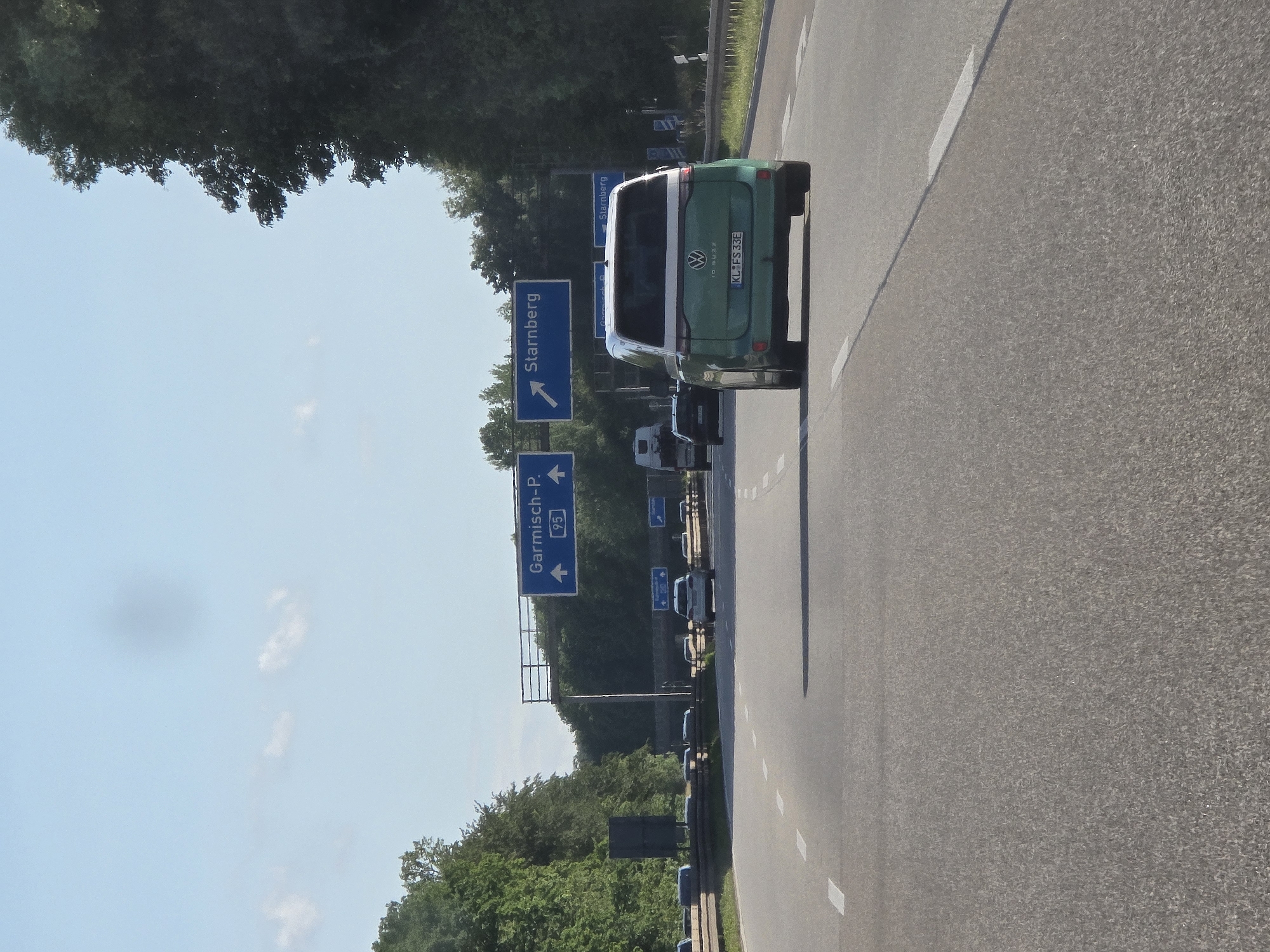 The image displays a daytime view of the German Autobahn A95, looking southbound towards Starnberg, Germany, on a clear day. The sky is light blue with a few scattered white clouds and a prominent dark, circular smudge in the upper center. Dense green deciduous trees line both sides of the multi-lane highway.
Overhead, a metal gantry supports two large blue directional signs. The left sign displays "Garmisch-P." (Garmisch-Partenkirchen) with an upward arrow and the Autobahn designation "95" in a white square. The right sign indicates "Starnberg" with an arrow pointing upward and slightly to the right, signifying an upcoming exit. Additional, smaller blue signs are partially visible below the main gantry.
The asphalt road surface features clear white dashed lane markings. A long, thin shadow, likely cast by the vehicle from which the photograph was taken, extends across the road from the lower right.
In the mid-ground, a two-tone green and white Volkswagen ID. Buzz electric van is proceeding in the rightmost lane. Its rear is visible, showing the VW logo, "ID. BUZZ" badging, and a German license plate reading "KL FS 33E". Behind the van, in the left lane, a dark-colored sedan with an obscured German license plate is visible. Further back, a white truck and several other indistinct vehicles are present on the highway and what appears to be a diverging lane or exit ramp on the left. No people are distinctly visible.