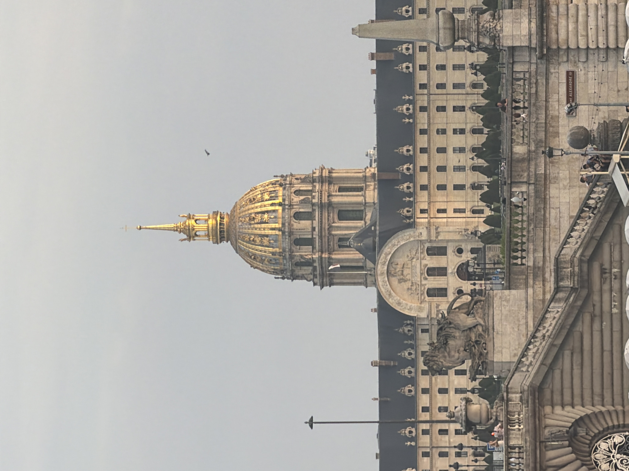 The image shows a view of the Dôme des Invalides in Paris, France, from a distance across the Seine River. The iconic golden dome is the central focus, bathed in what appears to be late afternoon or early morning light, highlighting its ornate details and the cross at its apex. Below the dome, the grand structure of Les Invalides stretches across the frame, a long, symmetrical building with numerous windows and architectural embellishments.
In the foreground, a stone bridge or embankment is visible, featuring decorative stonework, balustrades, and staircases. A prominent stone sculpture of a lion is positioned on the left side of the embankment. Several people are scattered across the bridge, some standing near the balustrade and others walking. Streetlights with spherical tops are also visible along the embankment. In the background, beyond the main building, more Parisian architecture can be seen, and a single bird is flying in the pale sky. The overall scene conveys a sense of historical grandeur and urban life in Paris.