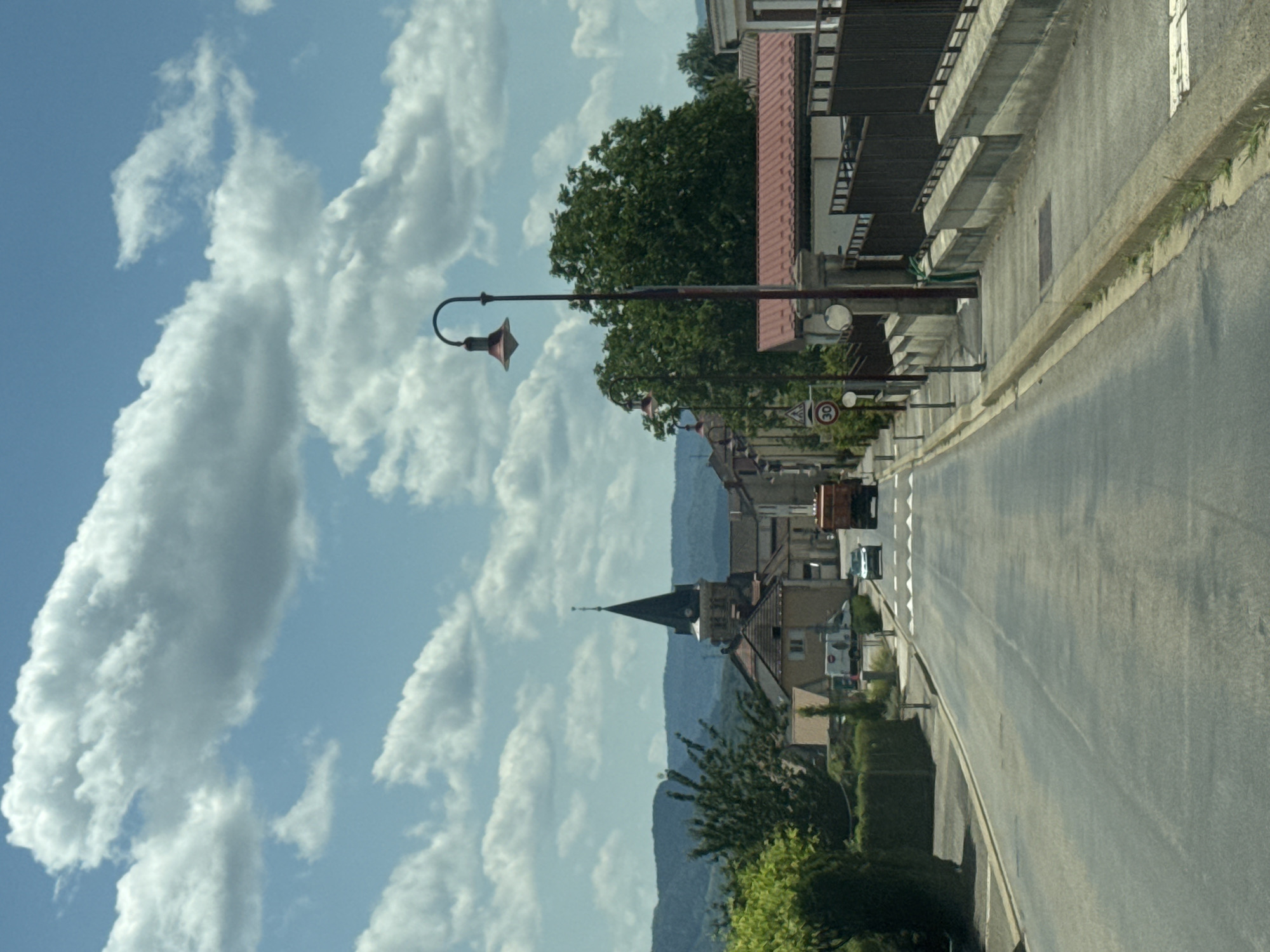 This is a street-level, eye-level photograph taken on a sunny day in Coteaux du Lizon, France. The image shows a two-lane asphalt road extending into the distance, with a sidewalk on the right side. The road is lined with trees and buildings, including a church with a tall steeple. A street lamp is prominently featured on the right side of the road. Several vehicles are visible on the road, including a car and a truck. The sky is blue with scattered white clouds. The overall scene depicts a typical French village or small town.