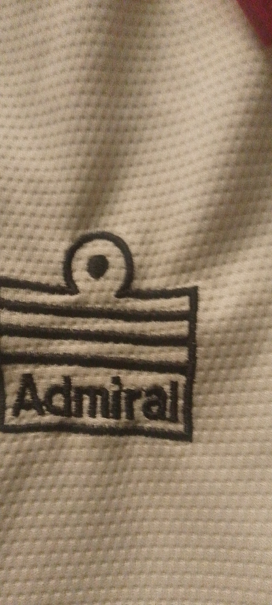 The image shows a close-up view of a beige fabric with a repeating dot pattern. A dark logo is embroidered on the fabric, featuring the word "Admiral" in capital letters at the bottom, within a rectangular outline. Above the word, there is a graphic element consisting of several horizontal lines and a circular shape with a dot in the center. The lighting in the image is somewhat uneven, casting shadows and creating highlights on the textured fabric. There is no discernible information about the location in Jalingo, Nigeria, or any other environmental context.