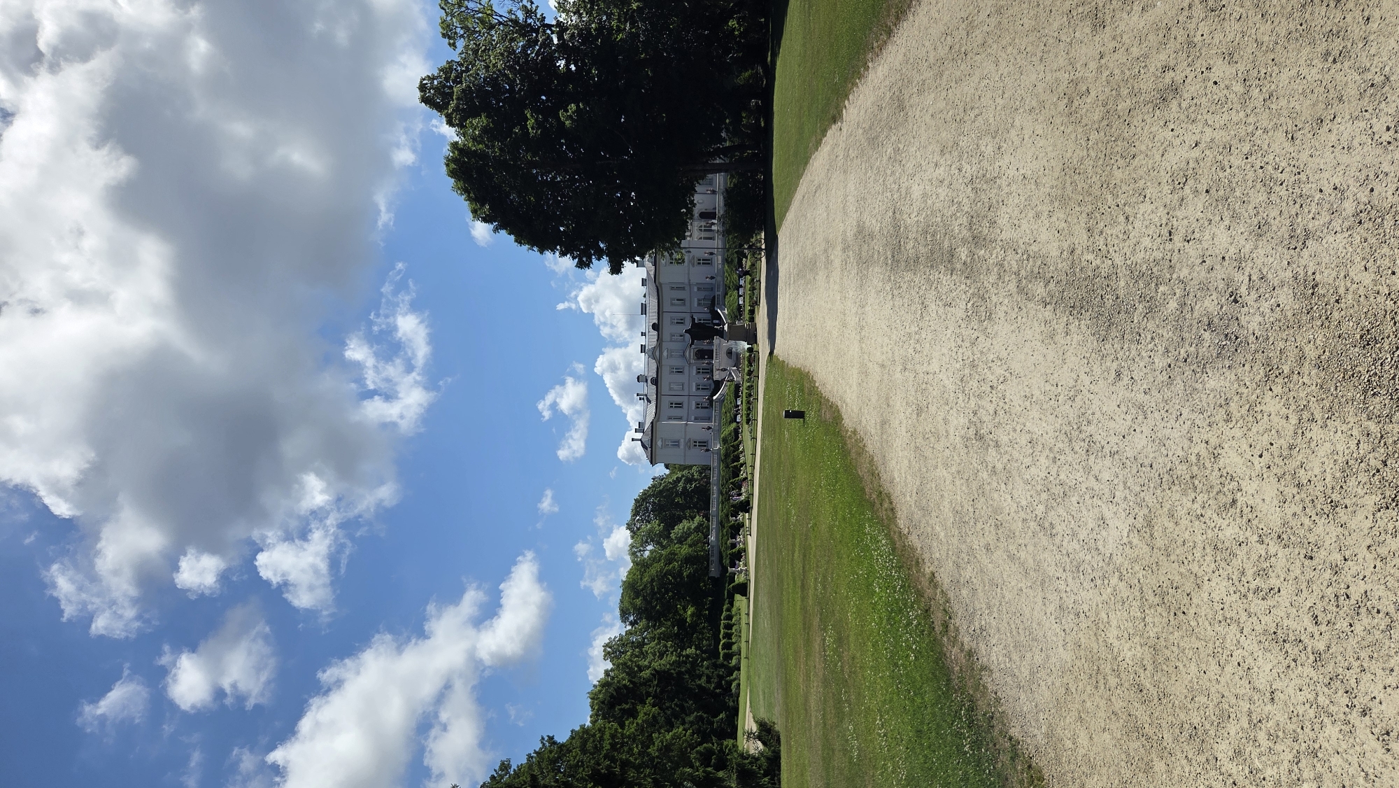 The image depicts a wide, gravel pathway leading towards a large, white manor-style building with a long facade and multiple windows. The building is situated amidst lush green grounds, with manicured hedges and scattered trees. A prominent dark-colored statue stands on a raised platform in front of the building, accessible by a set of stairs. The sky above is a clear blue, dotted with fluffy white clouds. The scene appears to be a well-maintained park or estate.
This location is the Palanga Amber Museum, also known as Tiškevičiai Palace, in Palanga, Lithuania.