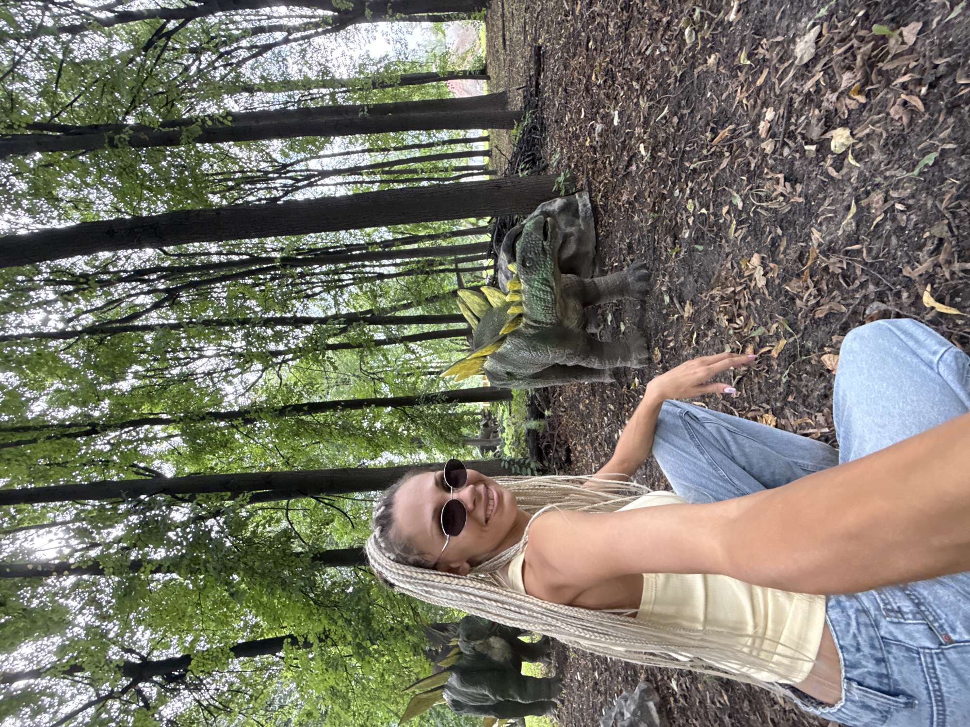 A young Caucasian woman with blonde braided hair, wearing a yellow tank top and blue jeans, is posing for a selfie in a wooded area. She is smiling at the camera and wearing sunglasses. In the background, two dinosaur replicas, likely Stegosaurus, are visible among the trees. The ground is covered with fallen leaves and dirt. The setting appears to be an outdoor park or exhibit. The image does not provide specific details about the city of Rusanovo, Russia.