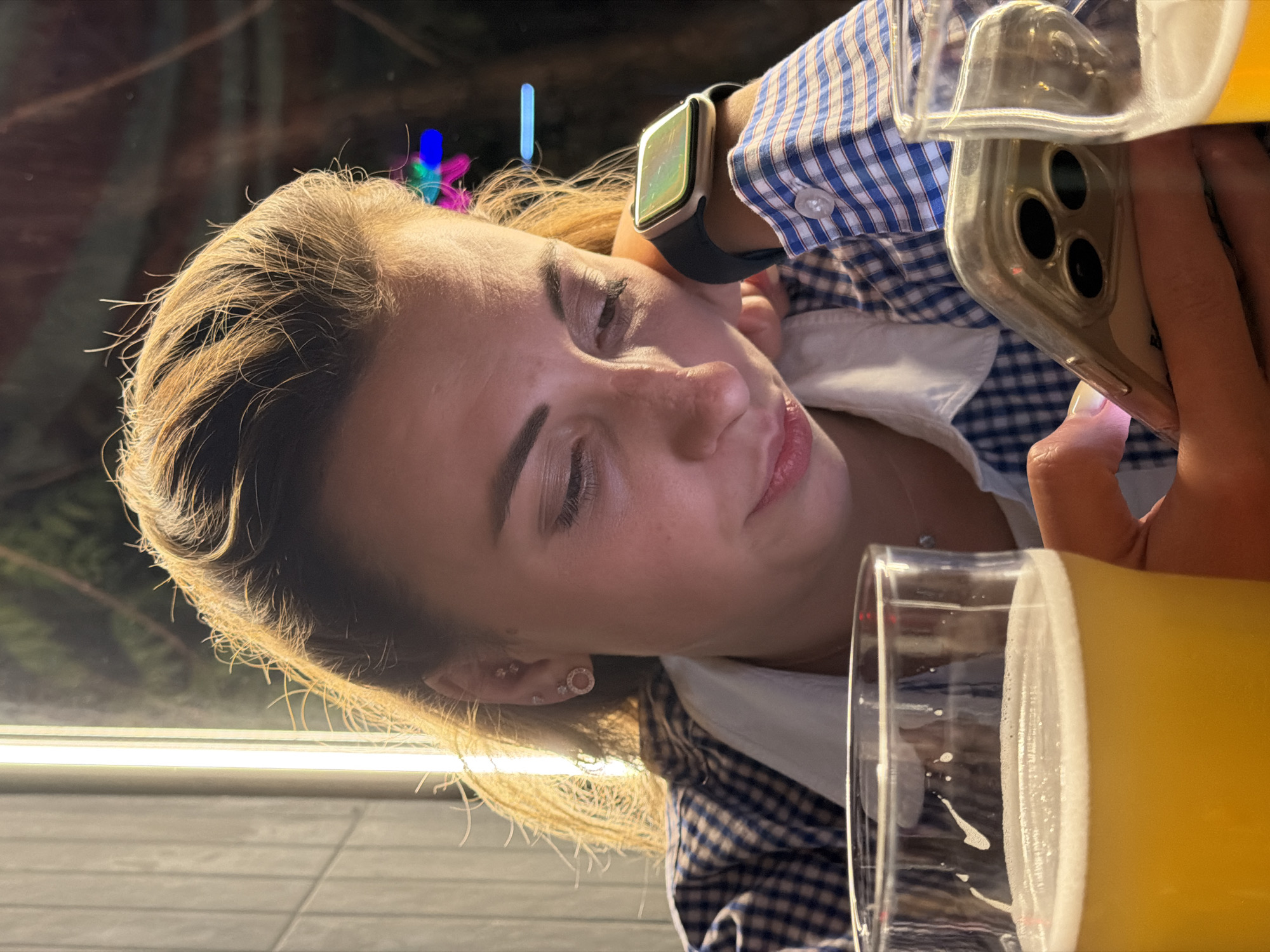 A woman with blonde hair is seen indoors in an undetermined location in Rostov-on-Don, Russia. She is wearing a blue and white checkered shirt over a white shirt. A smart watch is visible on her left wrist. The woman is looking at her phone, which has three camera lenses and is held in her right hand. A glass of beer with a foamy head is in the foreground. The background is dark with some light sources, including what appears to be neon lights.
