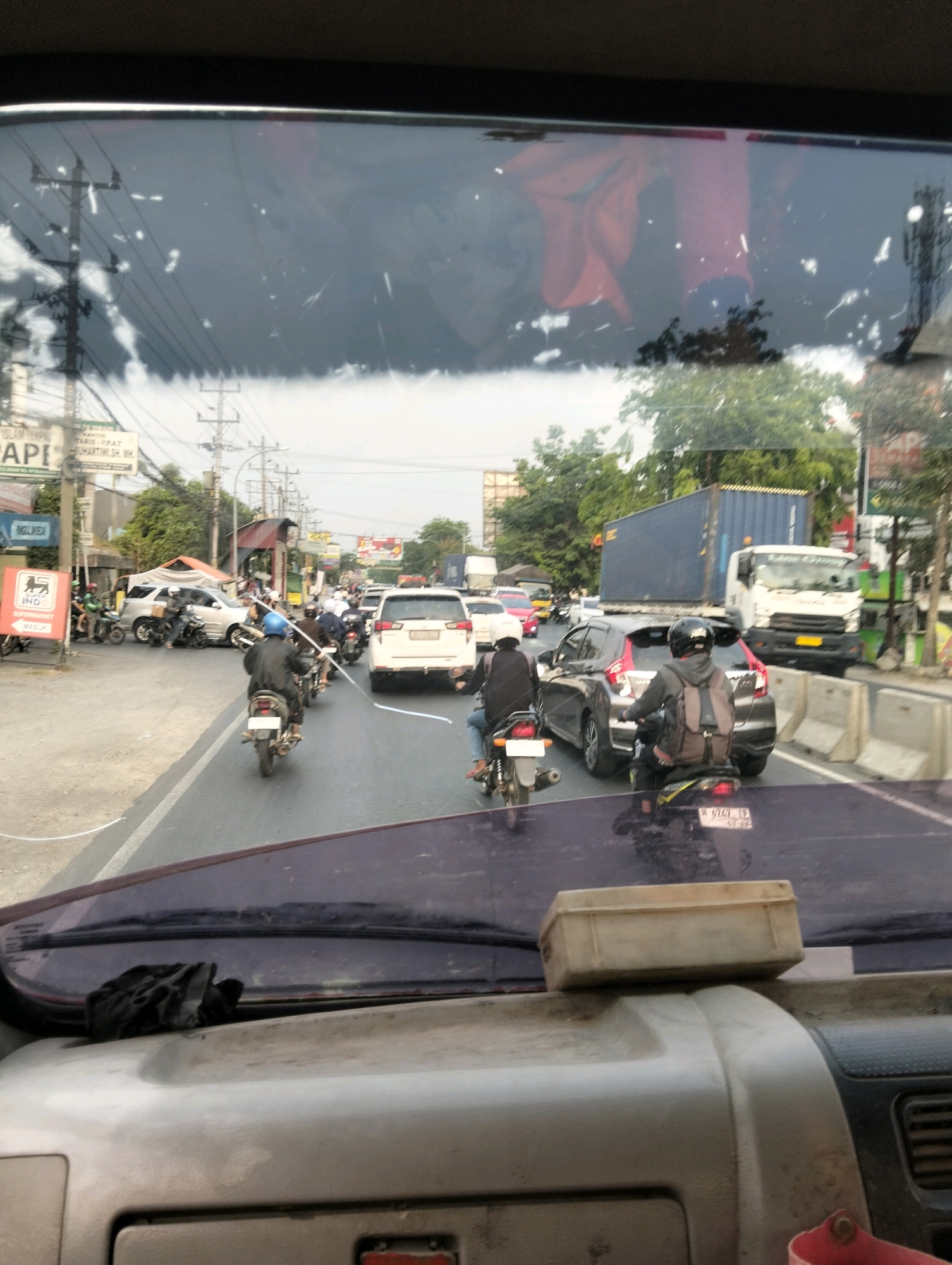 The image, taken from inside a vehicle, shows a congested road in Semarang, Indonesia. The foreground features the dashboard and windshield of the vehicle, with a dusty beige box resting on the dashboard. The windshield reflects the sky and surrounding environment, with streaks and smudges obscuring the view.
The road itself is filled with traffic, primarily motorcycles and cars. Several motorcycles are visible, with riders wearing helmets and backpacks. A white car is in the middle lane, and a dark-colored car is to its right. A white truck with a blue shipping container is stopped further down the road. Traffic lights and street poles line the sides of the road, along with buildings and trees. There is a sign for "SUPERMARKET MASUK" on the left side of the road. The overall scene depicts a typical busy street in Semarang, Indonesia, during daytime.