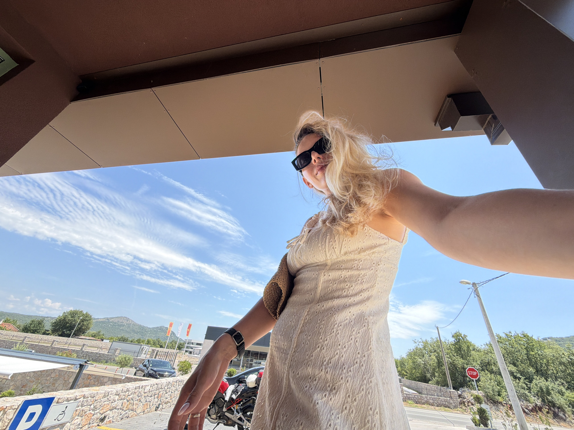 In Bigor, Montenegro, a woman is captured in a low-angle, wide-shot selfie. She is positioned centrally in the frame, wearing a white eyelet dress and black sunglasses, with her blonde hair windswept. She holds the camera out in front of her, her right arm extended. Her left arm is bent, with her hand resting near a wristwatch. A straw-colored item, possibly a bag or accessory, is slung over her left shoulder.
Behind her, a bright blue sky with wispy clouds dominates the upper half of the image. A brown overhang, part of a building's structure, frames the top. The lower half shows a street scene. A stone wall borders a parking area, marked with blue and white parking signs. A black car is parked to the left, and a motorcycle is visible to the right, partly obscured by the woman's arm. In the background, a road with a stop sign and telephone poles can be seen, with lush green trees and hills forming the distant landscape.