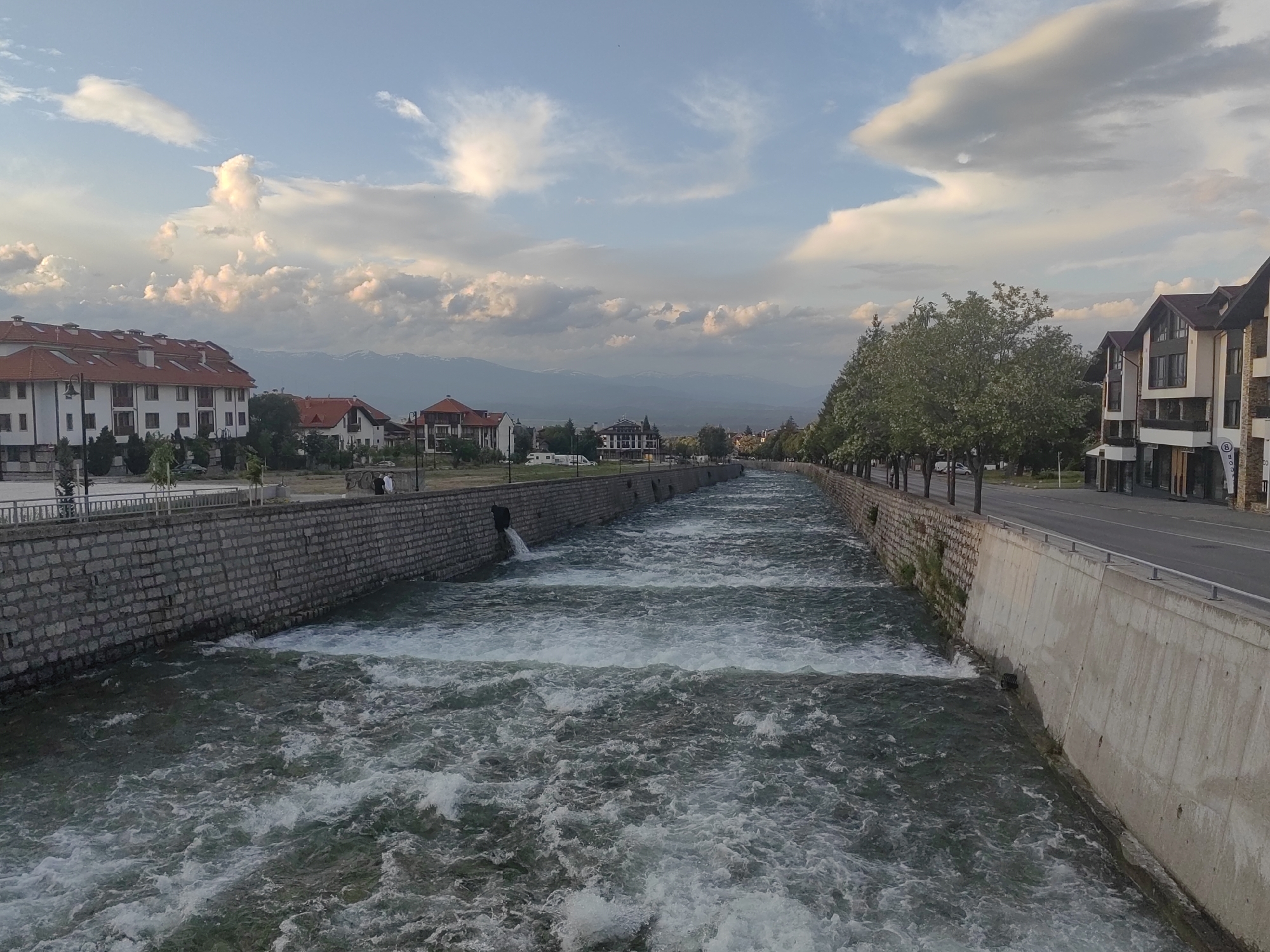 The media file displays a panoramic view of a wide, engineered river flowing through a populated area, with mountains in the background.
**Subjects:** The central subject is a river characterized by rapid, turbulent water, channeled between man-made embankments. The left bank features a stone wall, behind which stand multi-story buildings with red-tiled roofs and light-colored facades. Trees and streetlights line a paved area along this bank. On the right bank, a smooth concrete wall borders a paved road, which is also lined with numerous trees. Beyond the road, more multi-story buildings with darker roofs and modern designs are visible. In the distant background, a range of mountains, some with visible snowcaps, stretches across the horizon. The sky is partly cloudy with patches of blue, and scattered cumulus clouds. A few indistinct human figures can be seen on the left bank near the buildings.
**Activity or event taking place:** The image captures a static scene of an urban or semi-urban landscape. The primary "activity" is the natural flow of the river, which exhibits several small waterfalls or weirs, creating white water and turbulence. There are no overt human activities or events taking place, though the presence of buildings and roads indicates daily life in the area.
**Notable details:** The time of day appears to be late afternoon or early evening, indicated by the soft, slightly warm light on the clouds and structures. The weather is clear with scattered, well-defined clouds. The river's embankments are distinctly different on either side: stone on the left, concrete on the right. A pipe on the left stone wall is seen discharging water into the river. The architecture of the buildings on both sides suggests a European style.
**Visible text:** On a sign on the bottom-right building on the right bank, a faint and partially obscured word beginning with "BOG..." is visible. Another small, blue circular sign with unreadable white text is present on a building on the far right.