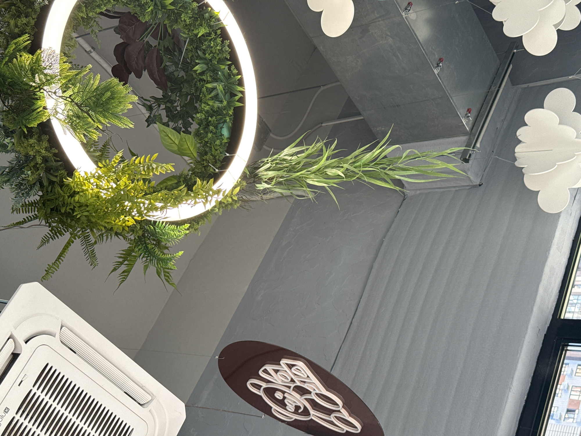 The image shows an interior scene, likely a commercial space, with various decorative elements and building features. Prominently displayed is a circular light fixture adorned with lush green artificial plants, creating a whimsical, nature-inspired ambiance. Dangling from the fixture is a long cascade of thin, vibrant green foliage, adding a dramatic vertical element. To the left, a dark brown oval sign with a white neon outline of a teddy bear and text is suspended, suggesting a playful or child-oriented theme. Above, a white air conditioning unit is visible, indicating a climate-controlled environment. The walls are painted a muted gray with a textured finish, and some exposed ductwork is visible in the upper right, hinting at industrial or utilitarian design elements. White cloud-shaped decorations are also present, contributing to the whimsical decor. The overall impression is of a modern, stylized interior with a focus on creating an engaging and visually interesting atmosphere. There is no indication of the city of Lyubertsy, Russia, in the provided image.