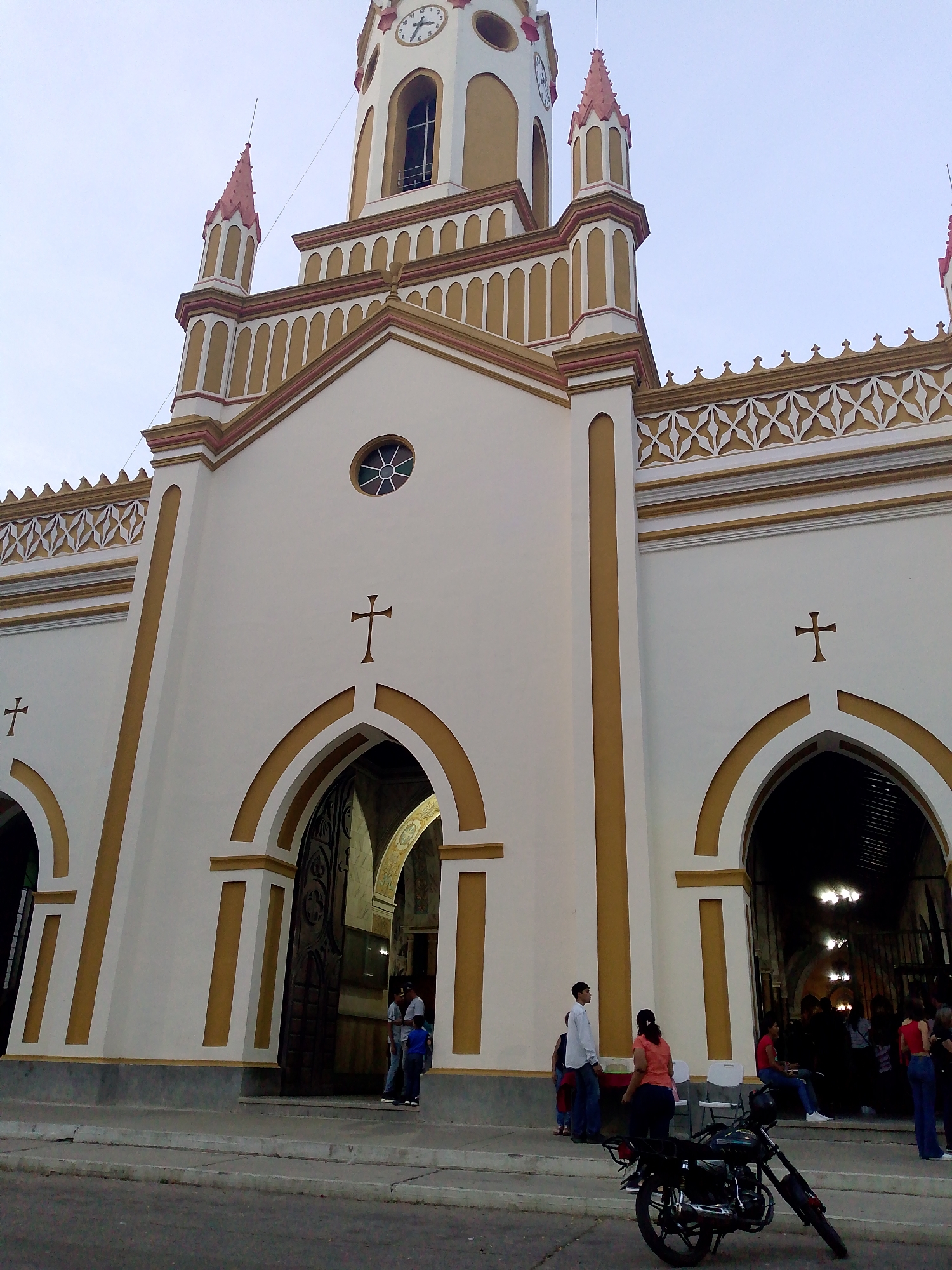 The image depicts the front facade of the church in Tinaquillo, Venezuela. The building is predominantly white with gold trim around the arches, columns, and roofline. The facade has three arched entrances, each topped with a cross. The central section of the facade has a round window above the central arch. The church has a central tower with a clock face and smaller towers on either side, decorated with red accents. Several people are standing near the entrances. A motorcycle is parked in front of the church. The sky is overcast.