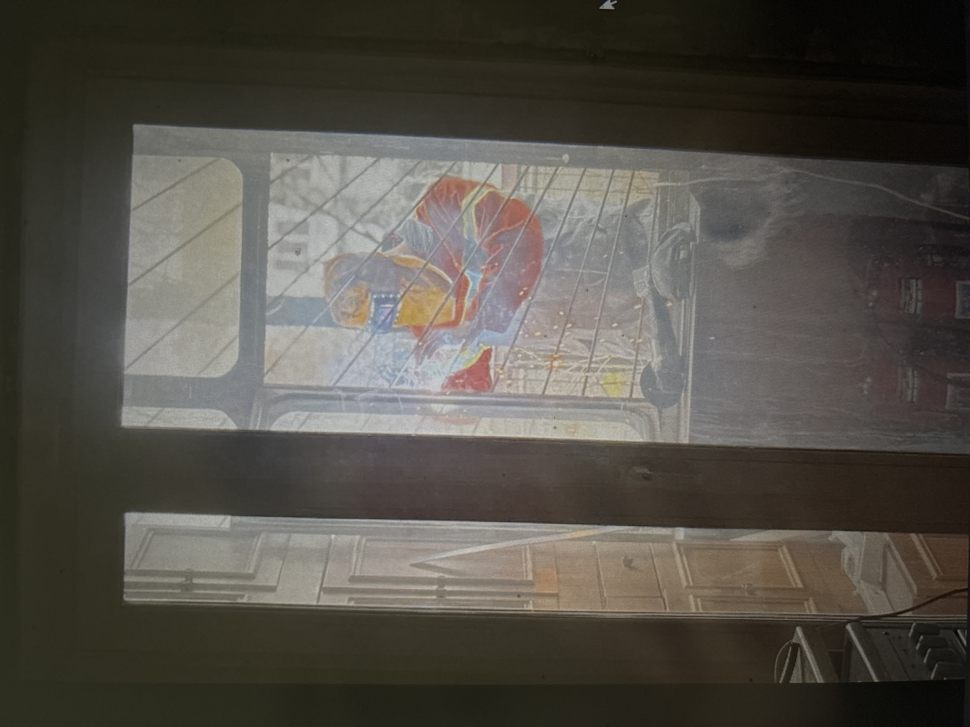 The image depicts a scene viewed through a window or glass door. Inside, a person wearing a welding mask and a red and yellow reflective jacket is welding. Sparks are visible from the welding activity. The background inside appears to be a cluttered room with shelves or cabinets. Outside the window, a metal grate or bars are visible, obscuring a full view of the exterior. Part of a wooden door frame is also prominent in the foreground. The overall lighting suggests an indoor environment with some natural light coming from the outside. There is no discernible indication of the specific location within Novosibirsk, Russia, beyond the provided context.