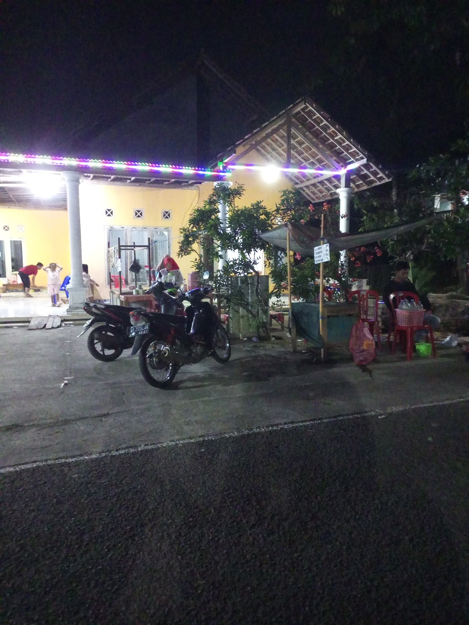 A nighttime outdoor scene in Jatipurno, Indonesia shows a yellow building with a covered porch illuminated by colorful string lights. Two motorcycles are parked on the asphalt in front of the building. A small roadside stall with a green tarp is set up under a canopy, with red chairs and a person sitting at a table. Several people are visible near the building entrance.