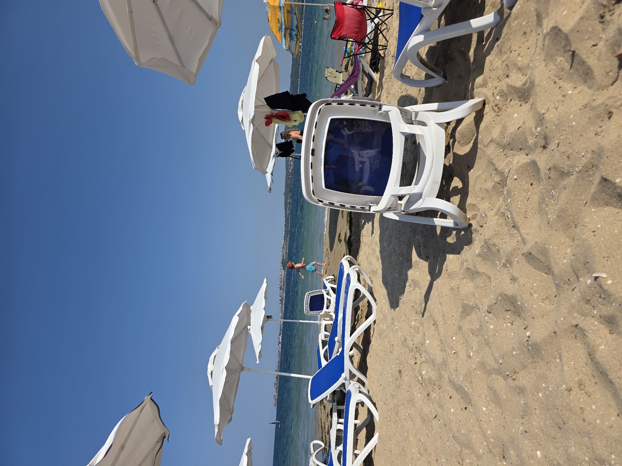 This is a medium-angle shot of a crowded beach on a sunny day, likely in Chernomorets, Bulgaria, with calm, blue waters meeting a sandy shoreline. In the foreground, several white plastic beach chairs with blue mesh seating are arranged on the sand, some angled towards the water. Behind these, rows of blue and white lounge chairs are set up, some occupied by people or covered by large white beach umbrellas. One white umbrella is prominent on the left, casting a shadow. Further back, the sea stretches out, with a distant shoreline featuring buildings and a clear blue sky above. A young child in blue shorts and a brown hat runs towards the water. In the mid-ground, several people are visible near the water's edge, some wading or swimming. One person is seated under a large white umbrella, with bags and clothing hanging from it. Another colorful umbrella with a tropical design is visible to the right, and further to the right, a red folding chair is set up. The sand is speckled with small shells and debris.