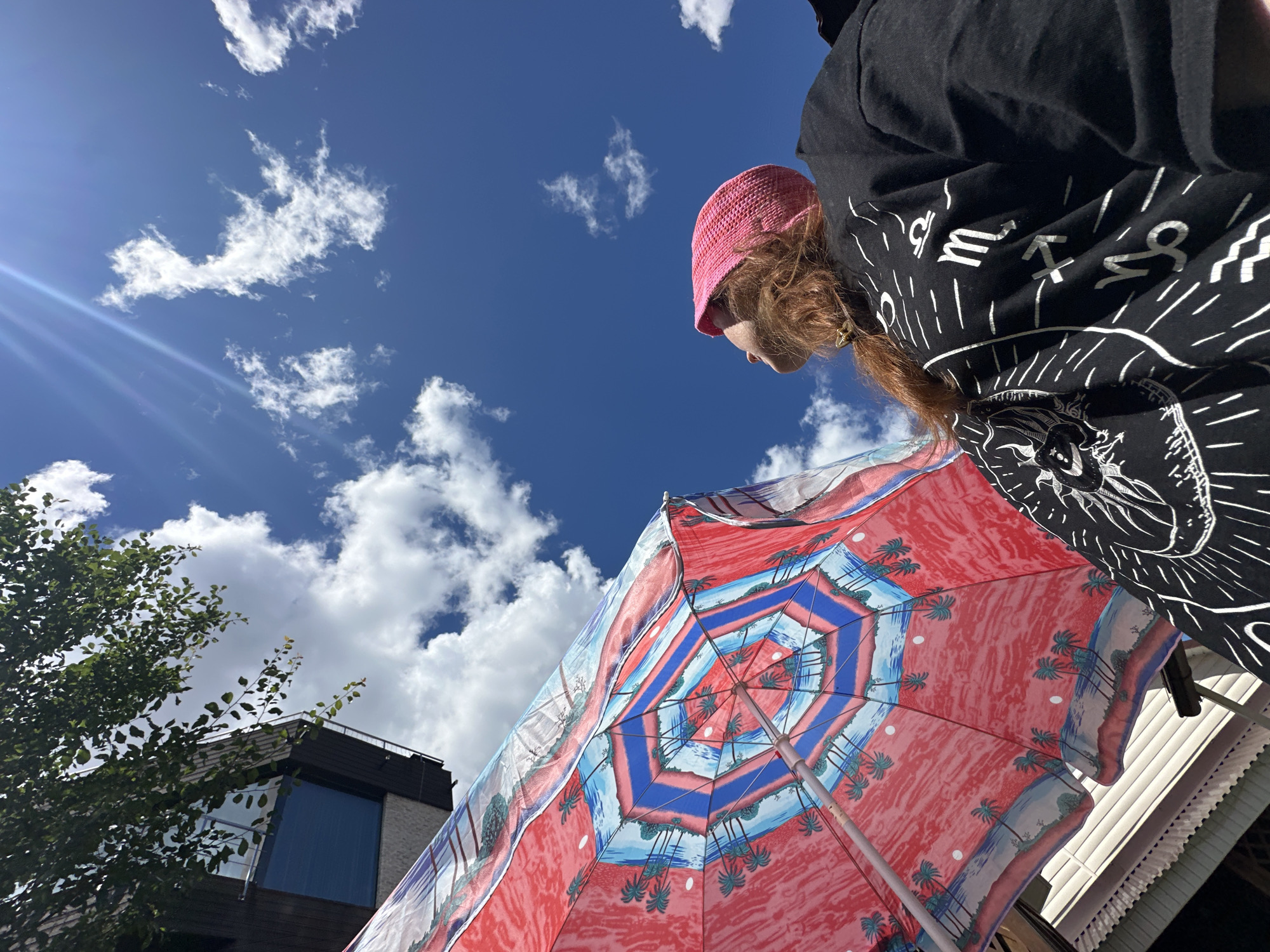A person with red hair and a pink knitted hat is looking up at a blue sky with white clouds. They are wearing a black t-shirt with white zodiac symbols. To the left is a red and blue umbrella with a tropical beach scene pattern. Part of a building and some green leaves are visible in the upper left corner. The sun is shining brightly, casting a lens flare. The image appears to have been taken outdoors in a city. There is no information in the image to determine the specific location in Nizhny Novgorod, Russia.