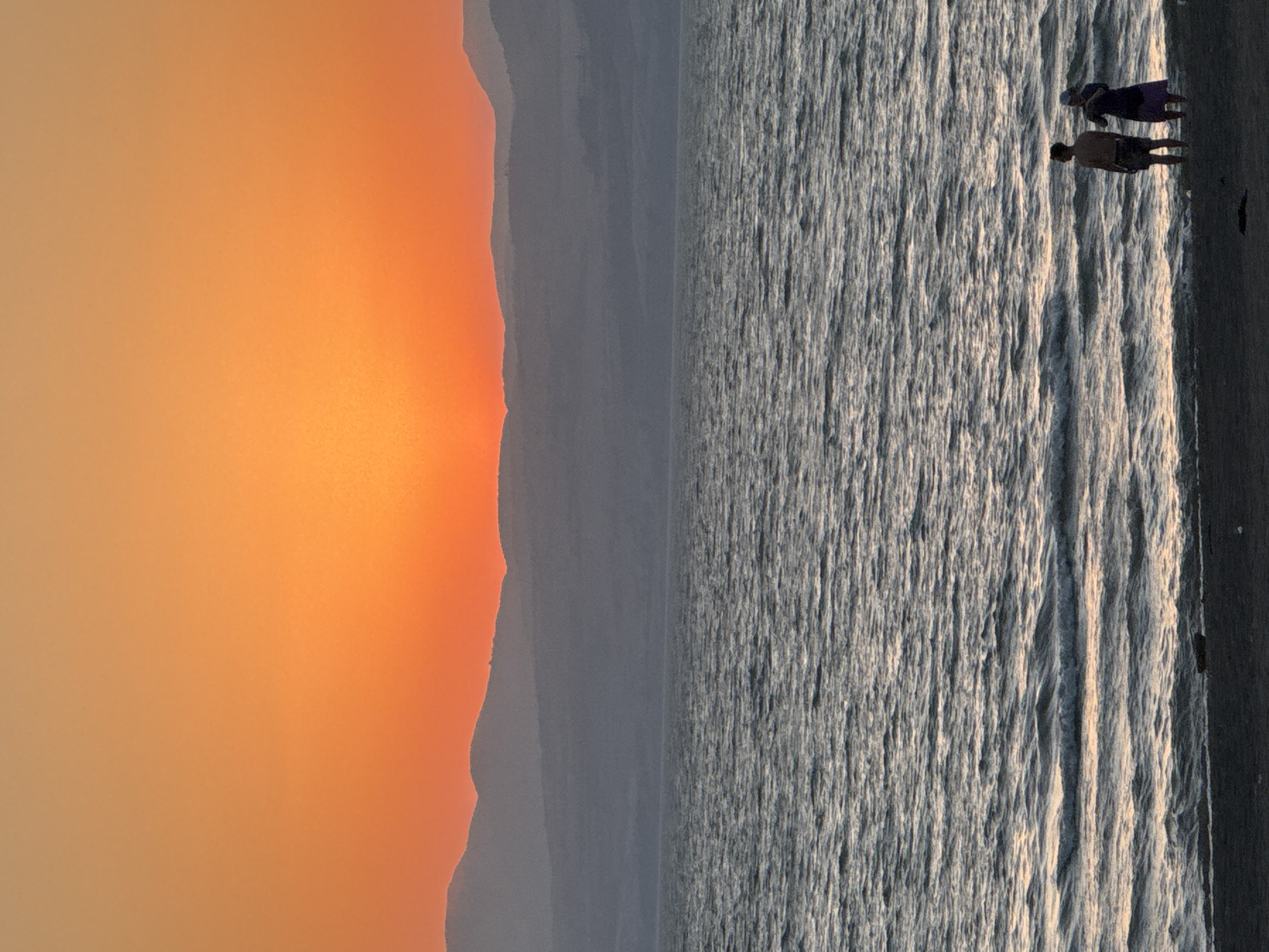 At dusk, two individuals stand on a sandy shore facing a body of water. The sky is a gradient of warm orange and yellow hues, with a subtle hazy mountain range in the background. The water exhibits a textured surface with small waves reflecting the sunset's glow. The individuals are silhouetted against the bright sky. The scene appears to be a serene moment at the Sea of Galilee in the Golan Regional Council, Israel.