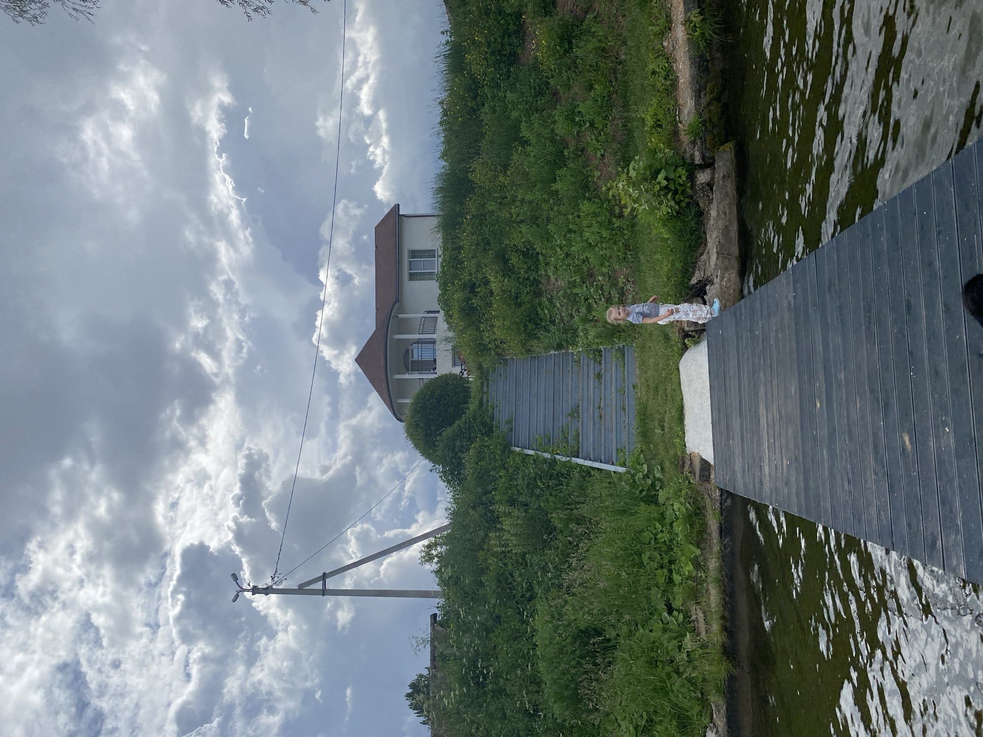 In Votkinsk, Russia, a girl stands on a wooden dock extending into dark, rippling water under a cloudy sky. The dock is dark-colored and made of planks. To the right of the girl, the dock meets the grassy bank. Behind the girl, a steep, overgrown green embankment rises, leading up to a white house with a red roof. A metal staircase is built into the embankment, ascending towards the house. A utility pole with wires stands to the left of the embankment. The overall scene suggests a rural or semi-rural setting by a body of water.