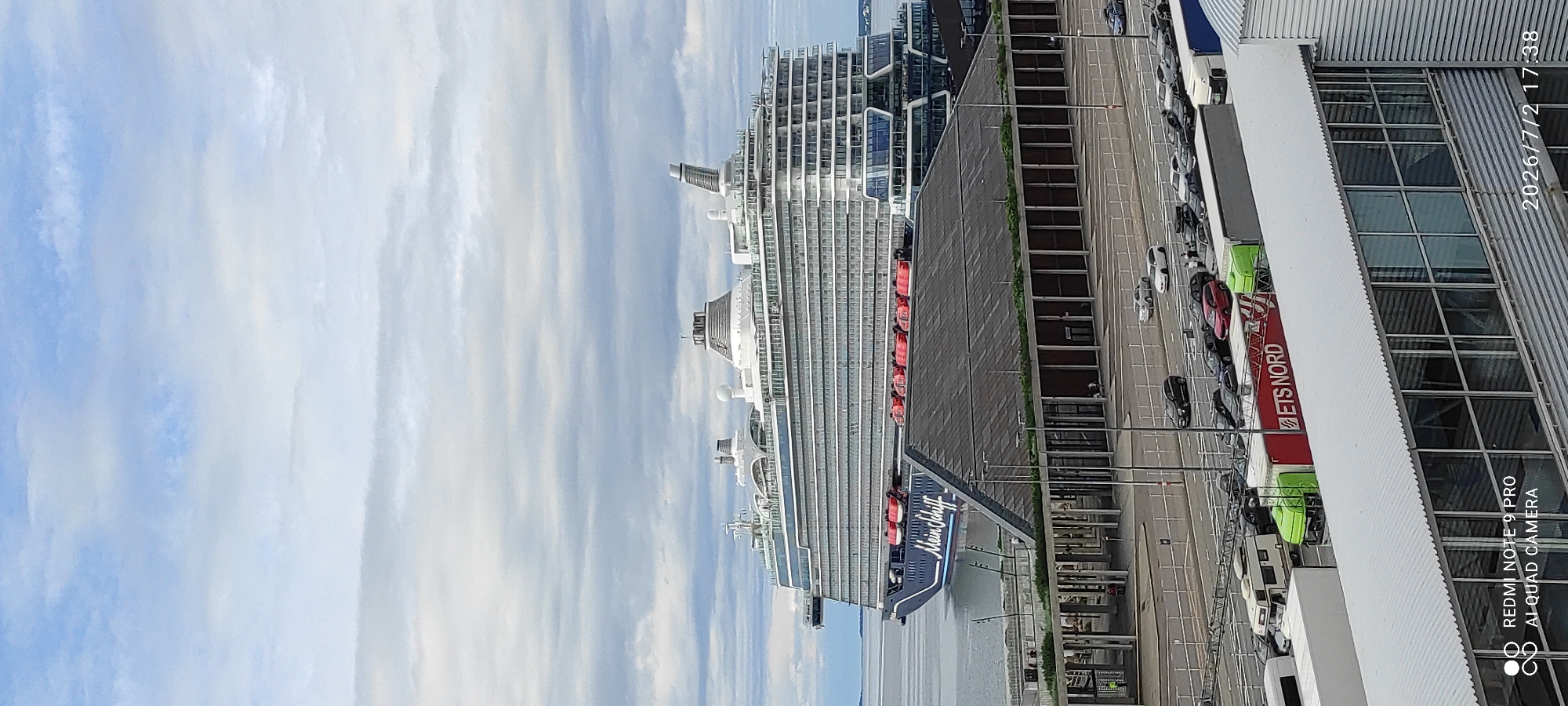 This is an aerial shot of a cruise ship docked at a port. The ship, named "Mein Schiff," is a large passenger vessel with multiple decks, white railings, and numerous lifeboats visible along its side. The port area features a long, dark-colored building with many windows, and a parking lot with several cars and trucks. One truck is prominently displaying "ETS NORD" in red lettering. The sky is filled with scattered clouds. The timestamp "2026/7/2 17:38" suggests the photo was taken in the late afternoon. The presence of the cruise ship and port infrastructure indicates a busy travel hub. The overall scene appears to be functional and organized, with no discernible emotions. The location context of Tallinn, Estonia, places this scene within a Baltic Sea port.